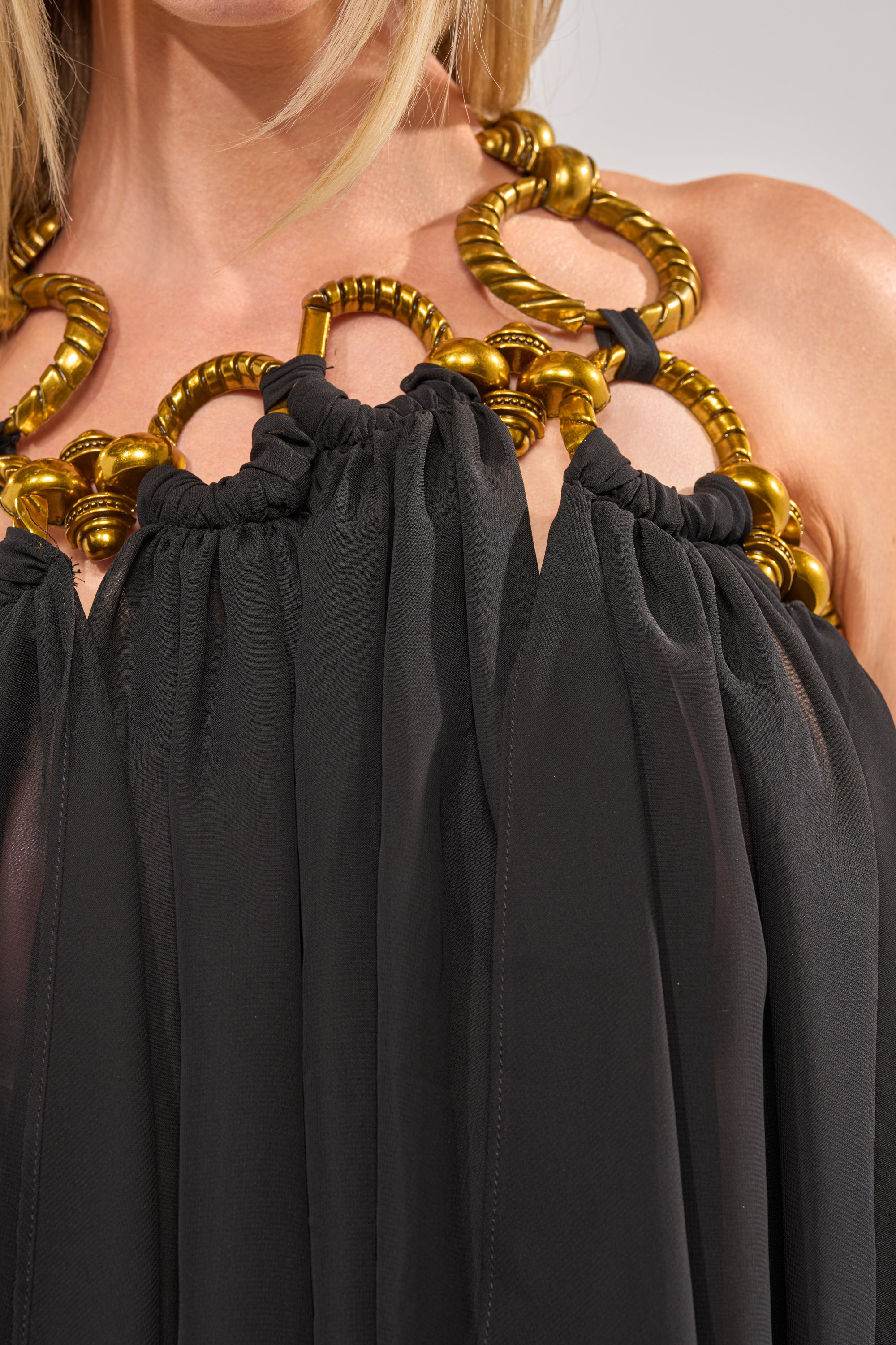 A close-up of a person wearing the DANCE THE NIGHT AWAY BLOUSE—black organza with large gold circular accents along the neckline, ideal for chic evening looks. Shoulders and part of the neck are visible. 7