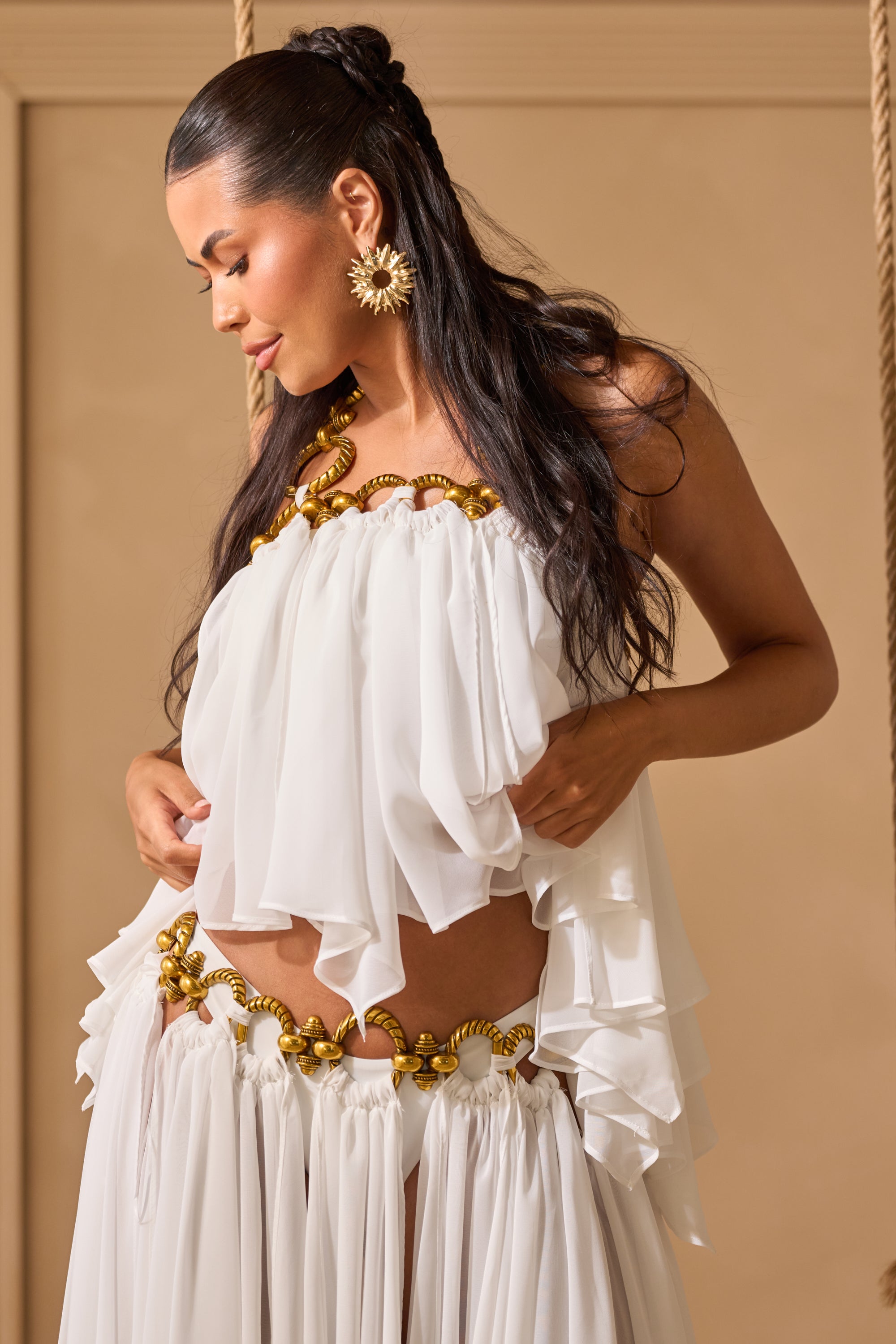 A woman with long dark hair and sun-shaped earrings gazes down, wearing the DESERT VIBES BRALETTE—a flowing white organza halter top with gold hardware—layered summer style against a soft beige background. 1