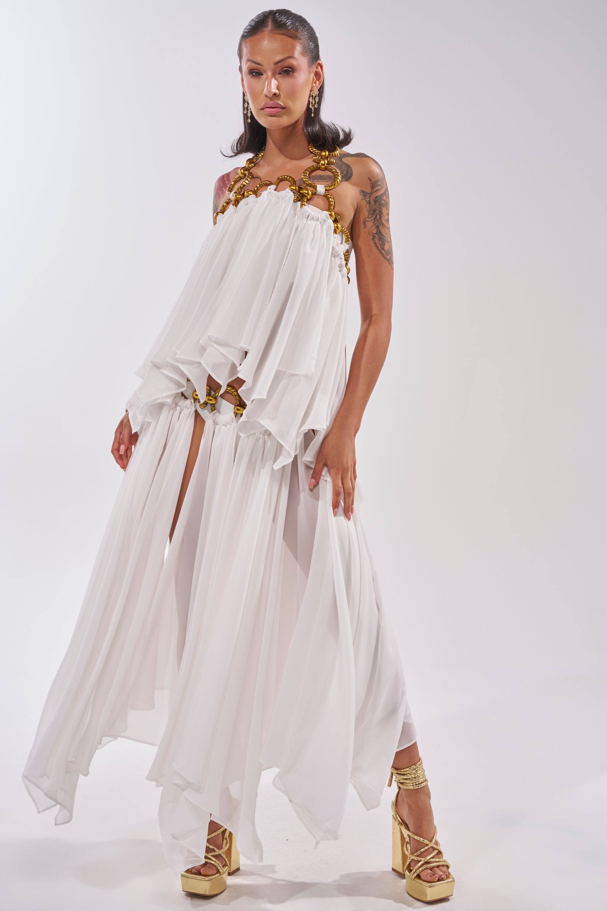A woman exudes confidence in the DESERT VIBES BRALETTE, styled with ruffled layers, high slits, gold hardware accents, and platform heels. Her arm tattoos show against a softly lit backdrop, evoking carefree summer vacation vibes. 5