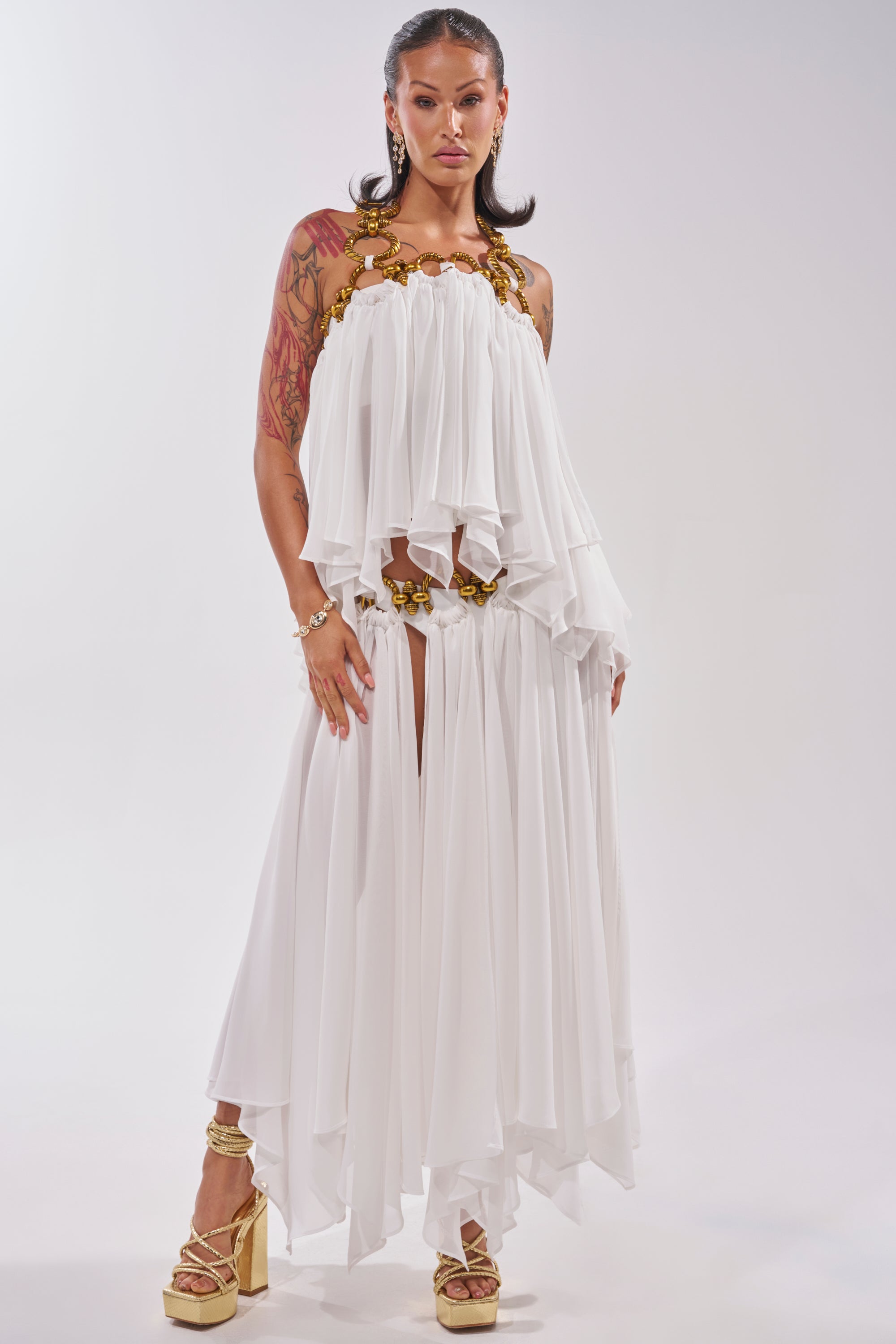 A woman exudes confidence in the DESERT VIBES BRALETTE, styled with gold heels and visible tattoos. Her dark hair is elegantly done, standing against a plain, light background. Gold hardware accents the look for a modern finish. 6