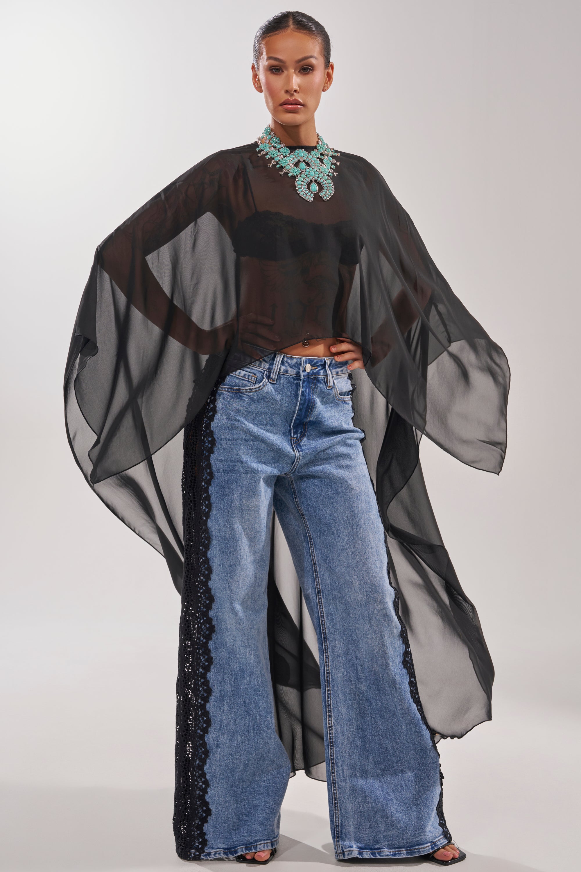 A woman poses in the COCO BLOUSE—a sheer chiffon top with cape sleeves—styled over a black bralette, wide-leg blue jeans with black lace sides, open-toe heels, and a bold turquoise necklace against a plain background. 0