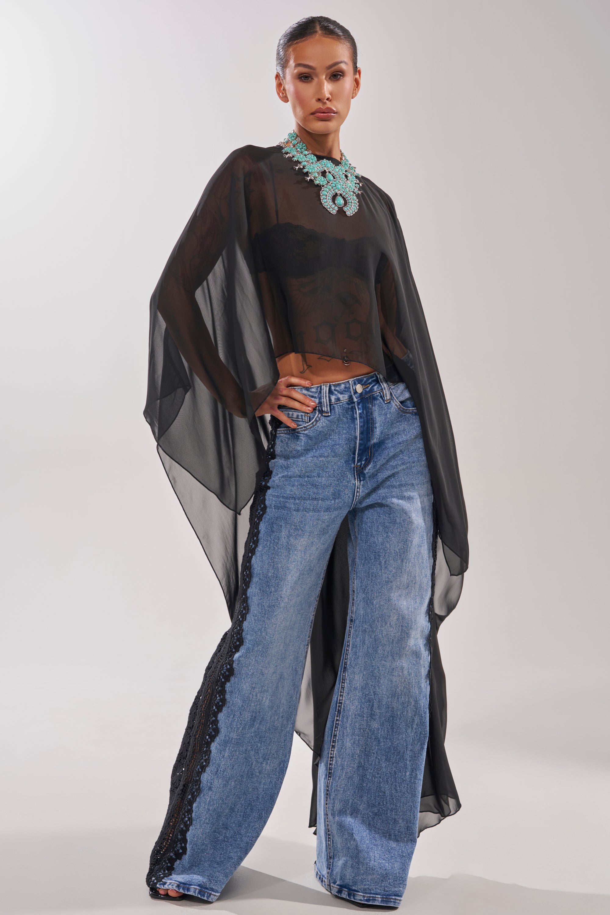 A woman poses confidently in the COCO BLOUSE, a sheer chiffon top with an asymmetrical hem over a black bralette, styled with wide-leg blue jeans and a bold turquoise necklace against a plain light background. 3
