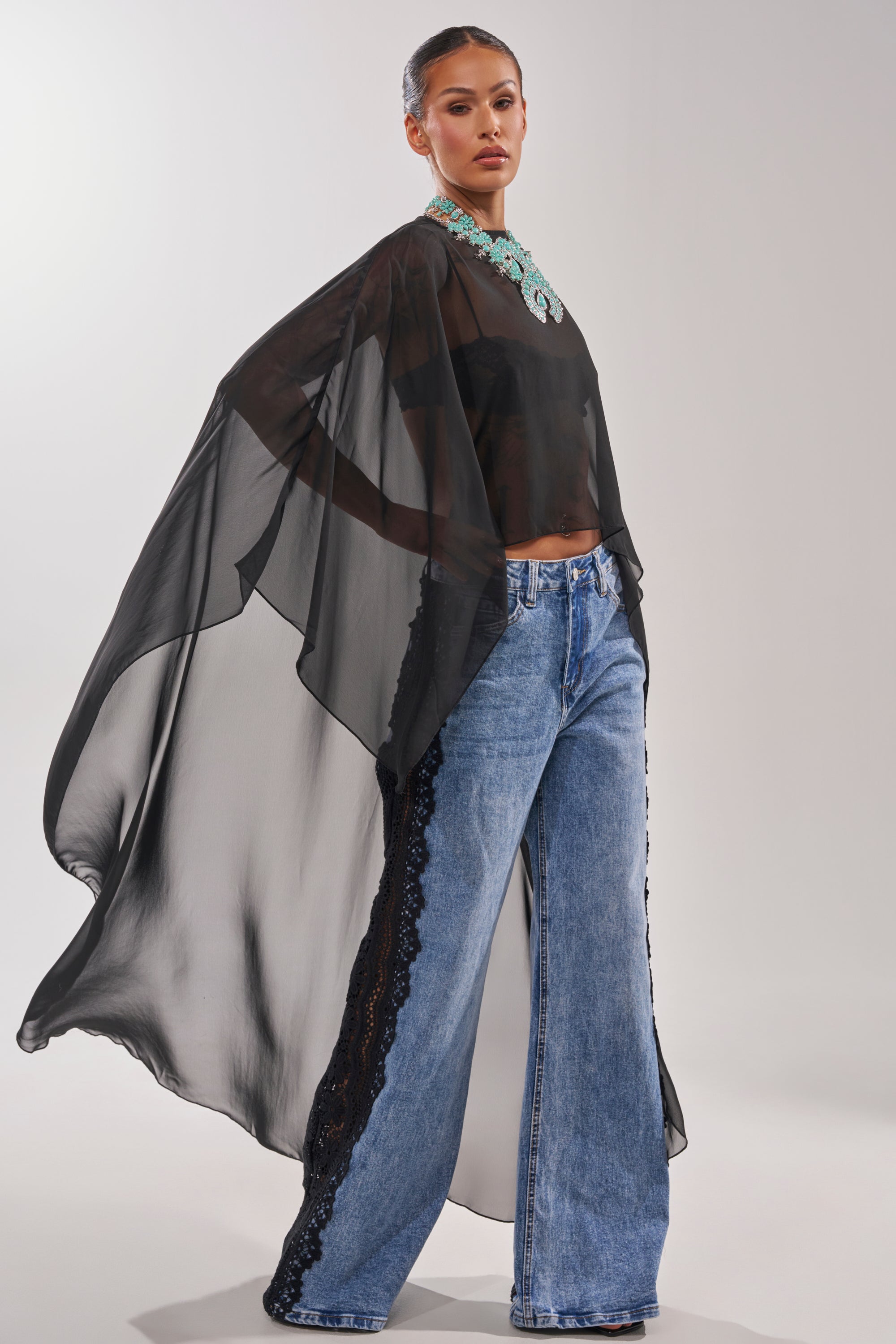A woman models the COCO BLOUSE—a sheer chiffon top with cape-style sleeves—paired with wide-leg blue jeans featuring black lace side panels and a statement turquoise necklace, all set against a plain light background. 4