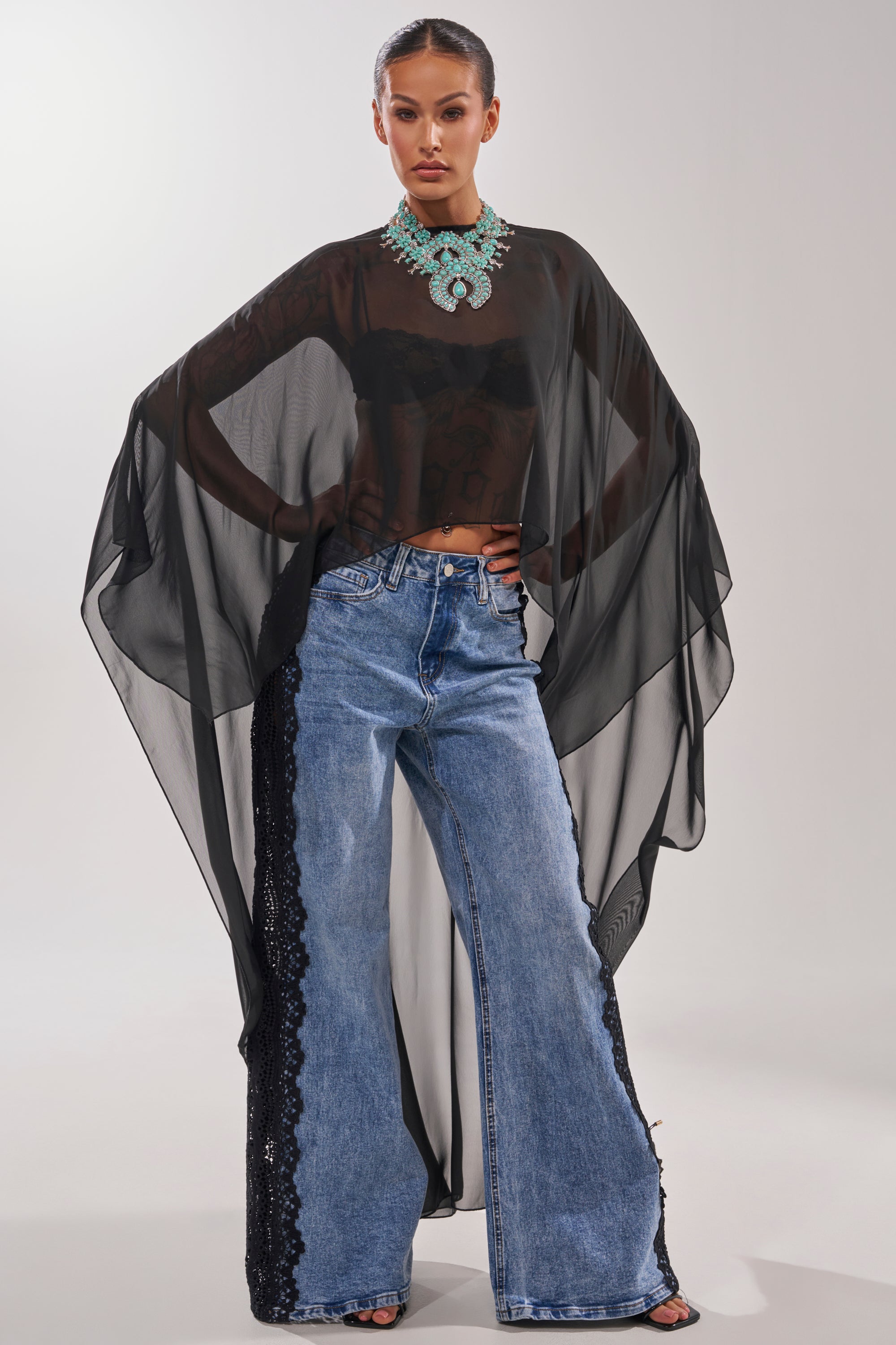 A woman poses confidently in the COCO BLOUSE, a sheer chiffon top with cape-style sleeves, styled with wide-leg blue jeans featuring black lace panels, a black bandeau, and a bold turquoise necklace against a plain light background. 5