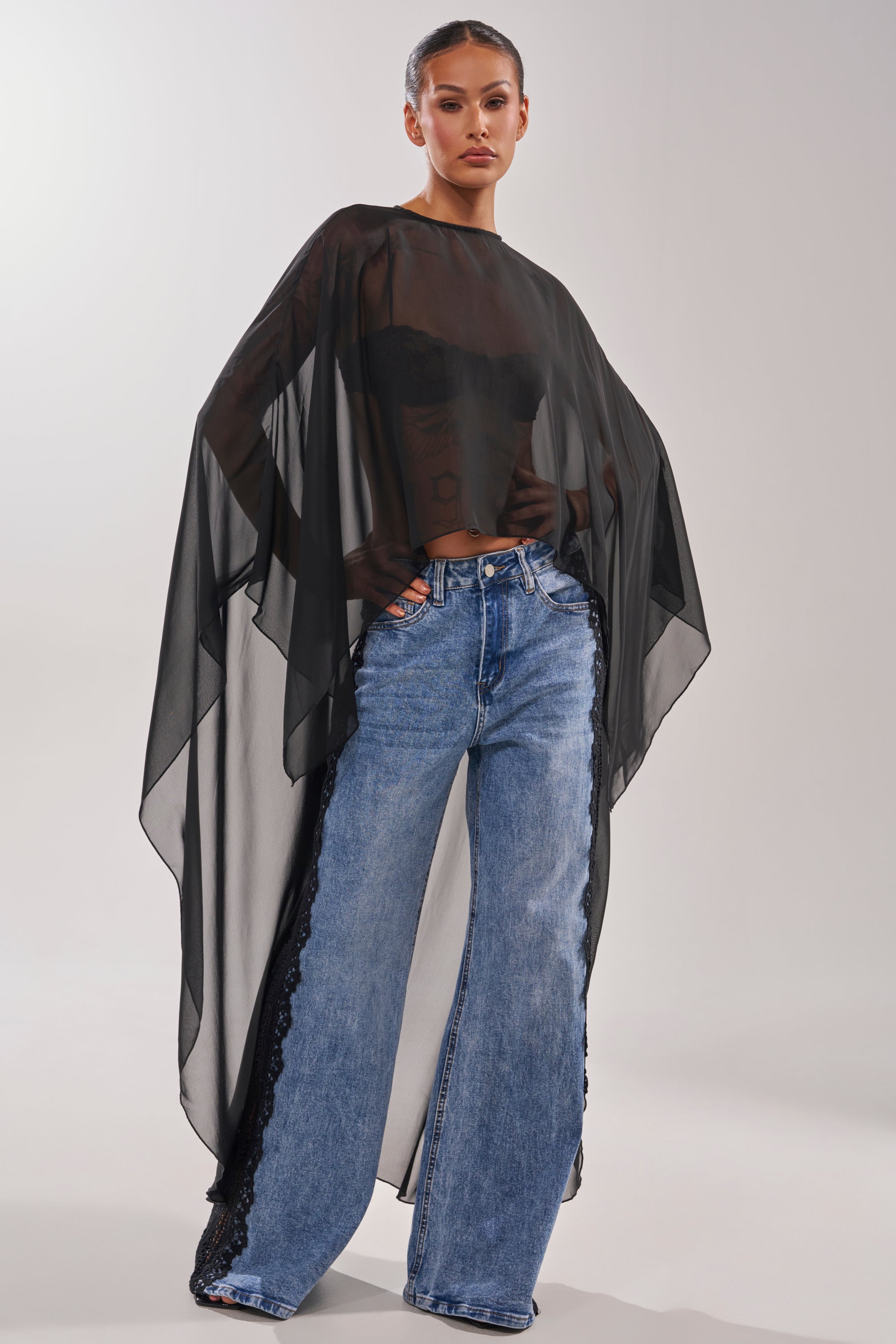 A person stands confidently in the COCO BLOUSE—featuring sheer chiffon, cape sleeves, and an asymmetrical hem—layered over a black corset and wide-leg blue jeans with lace details against a plain light background. 6