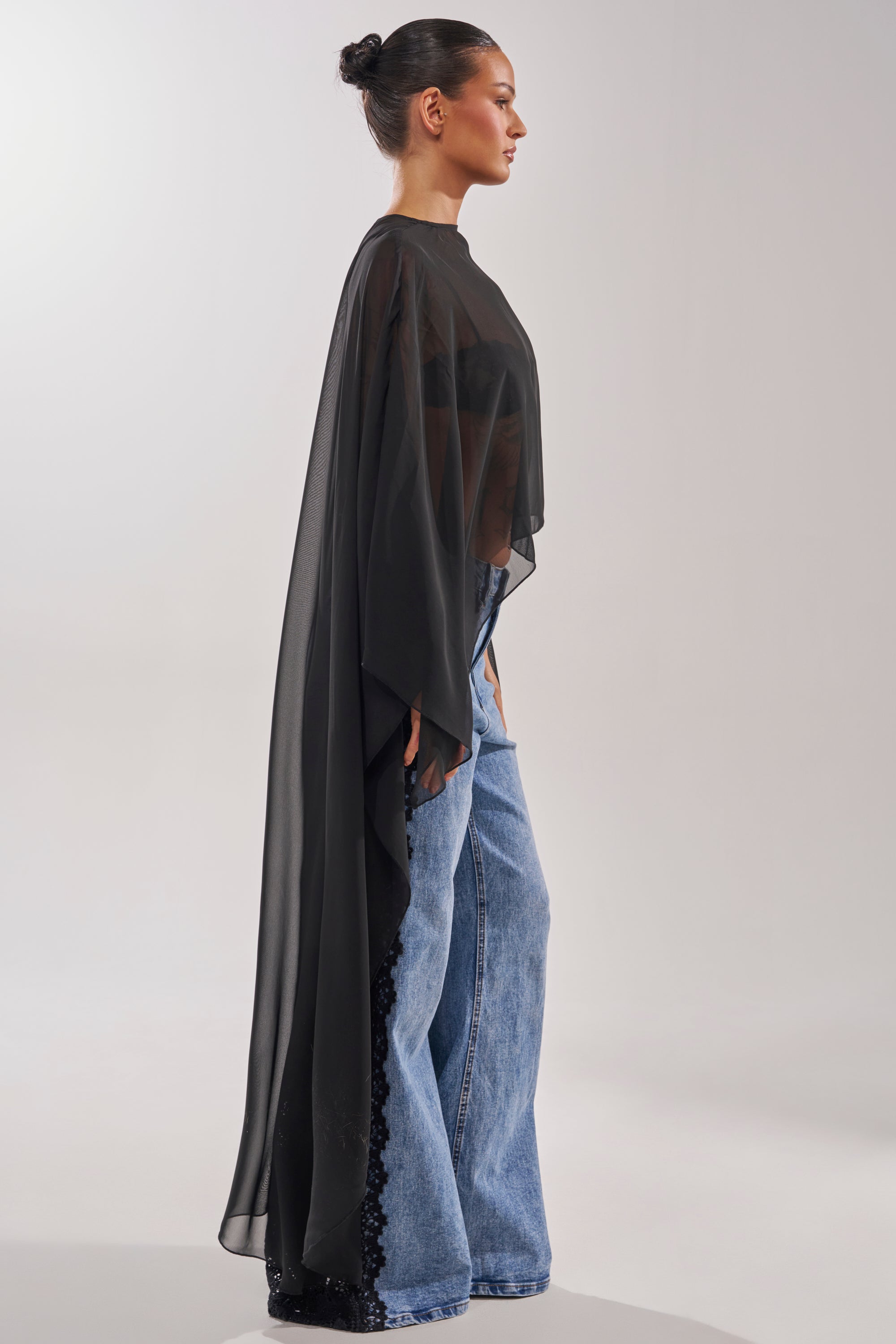 A woman stands in profile against a plain, light background, wearing wide-leg blue jeans and the COCO BLOUSE—a sheer chiffon top with cape-style sleeves and a floor-length asymmetrical hem. Her hair is styled in a sleek bun. 7