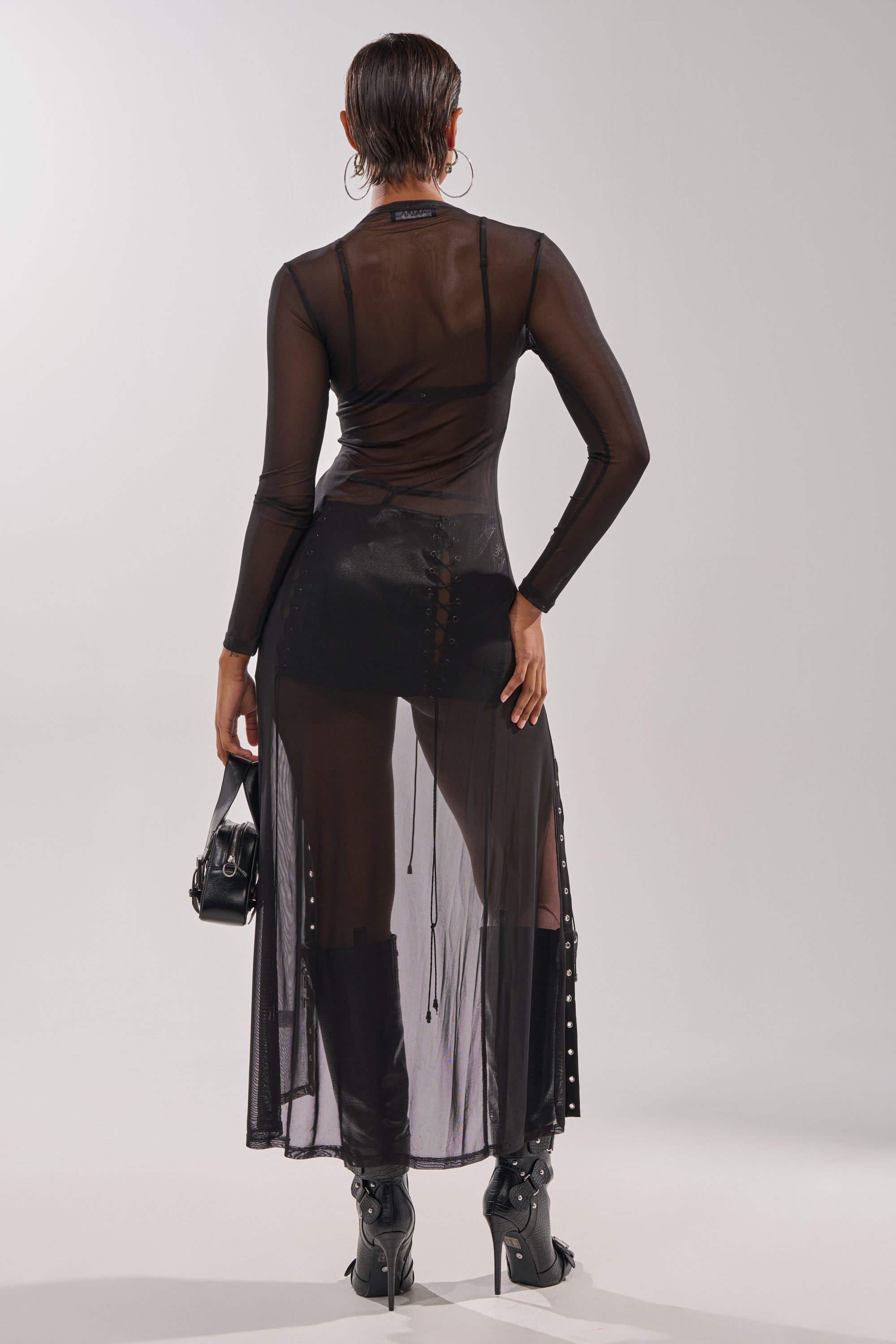 A person with wet-look hair, wearing the SEE THROUGH ME BLOUSE—a sheer black long-sleeve top with lace-up paneling and side slits—over black shorts and boots, stands facing away while holding a small black handbag. 3