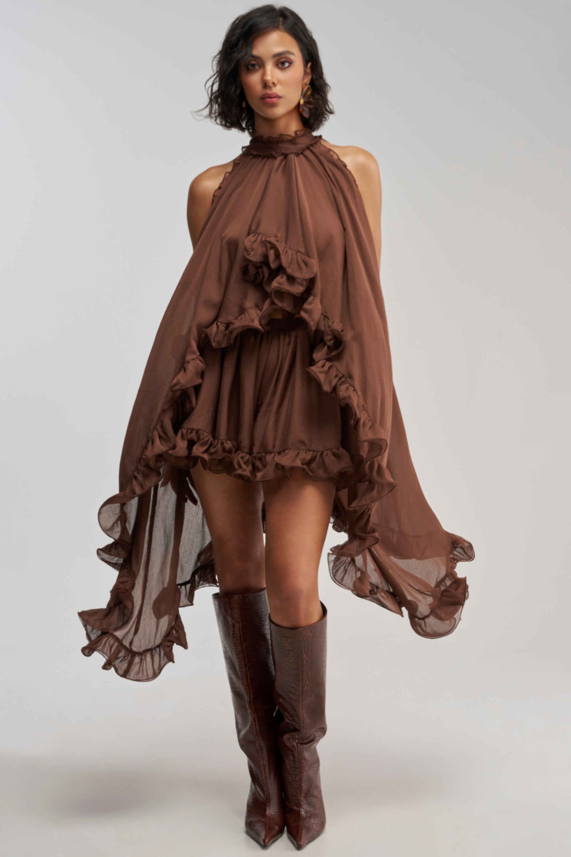 Wearing the TALK TO ME CHIFFON HIGH LOW BLOUSE—a brown chiffon top featuring ruffles, a high neckline, and a sleeveless design with a layered hem—paired with knee-high brown boots, the person stands against a plain light background.