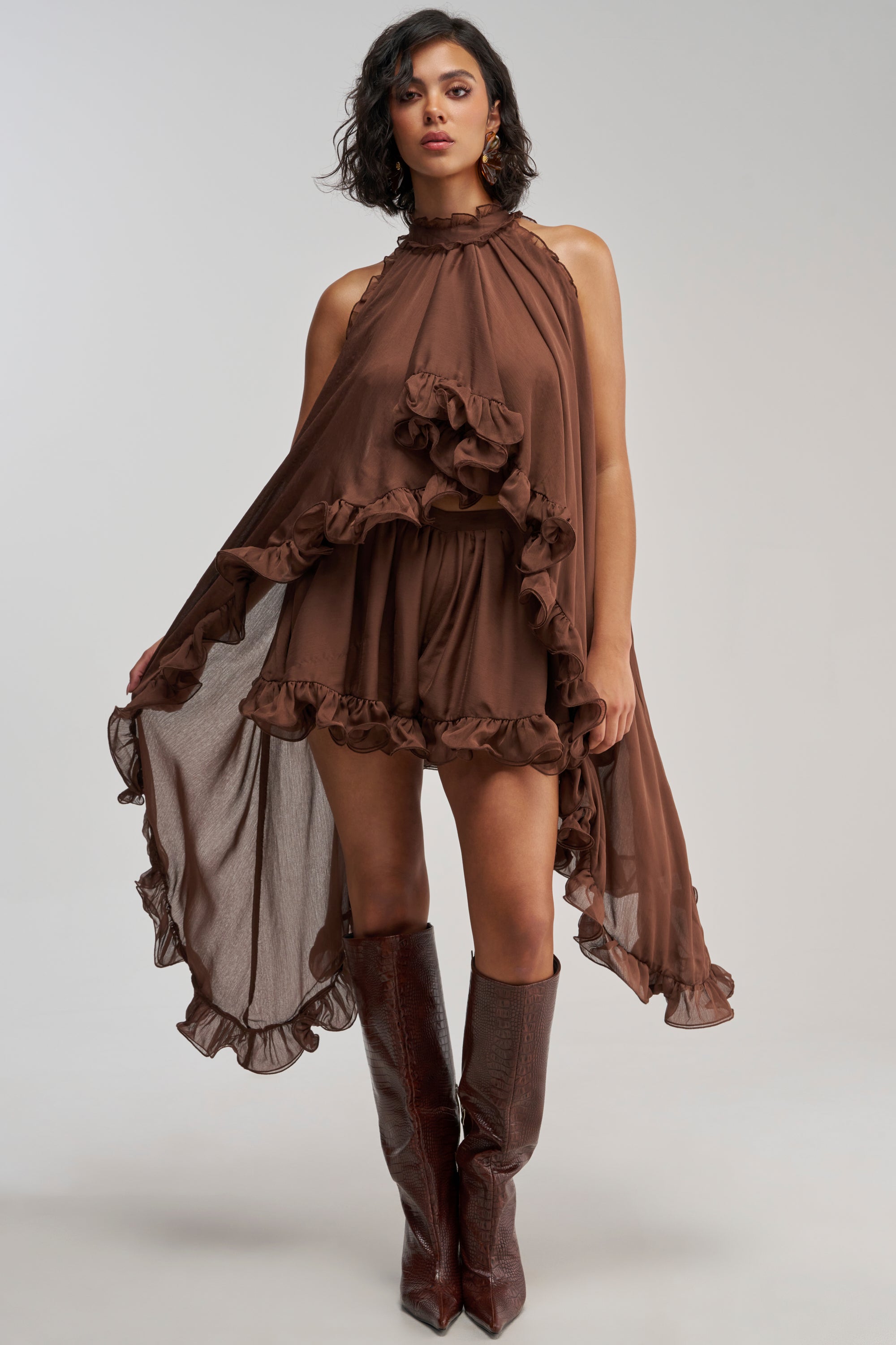 A woman wearing the TALK TO ME CHIFFON HIGH LOW BLOUSE—a sleeveless, high-neck, brown ruffled blouse with an asymmetrical hem—paired with matching brown knee-high boots stands facing forward.