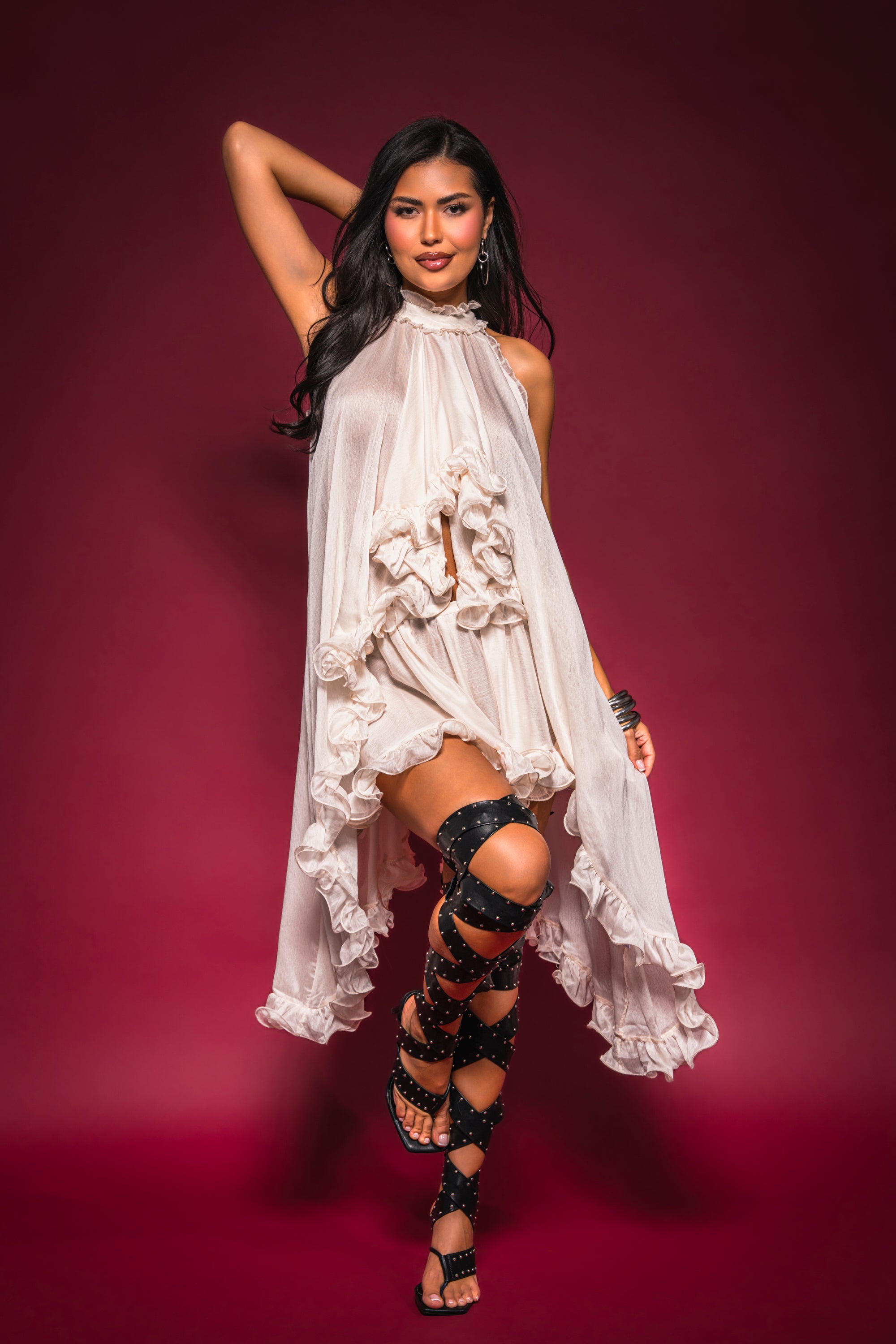 A woman poses confidently in the TALK TO ME HIGH LOW MESH BLOUSE, styled as a dress with a flowing hem and paired with black strappy knee-high sandals, set against a dark red background. 2