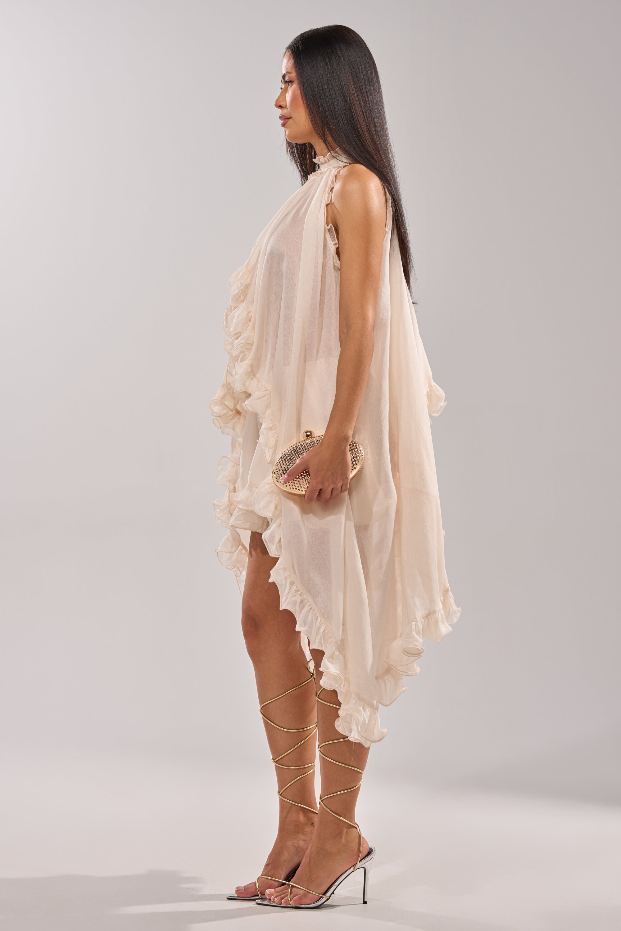 A woman in profile wears the TALK TO ME HIGH LOW MESH BLOUSE—a cream high-low blouse with ruffles—paired with strappy heels and a textured clutch. Her long, straight hair falls gracefully against a plain, light background. 6