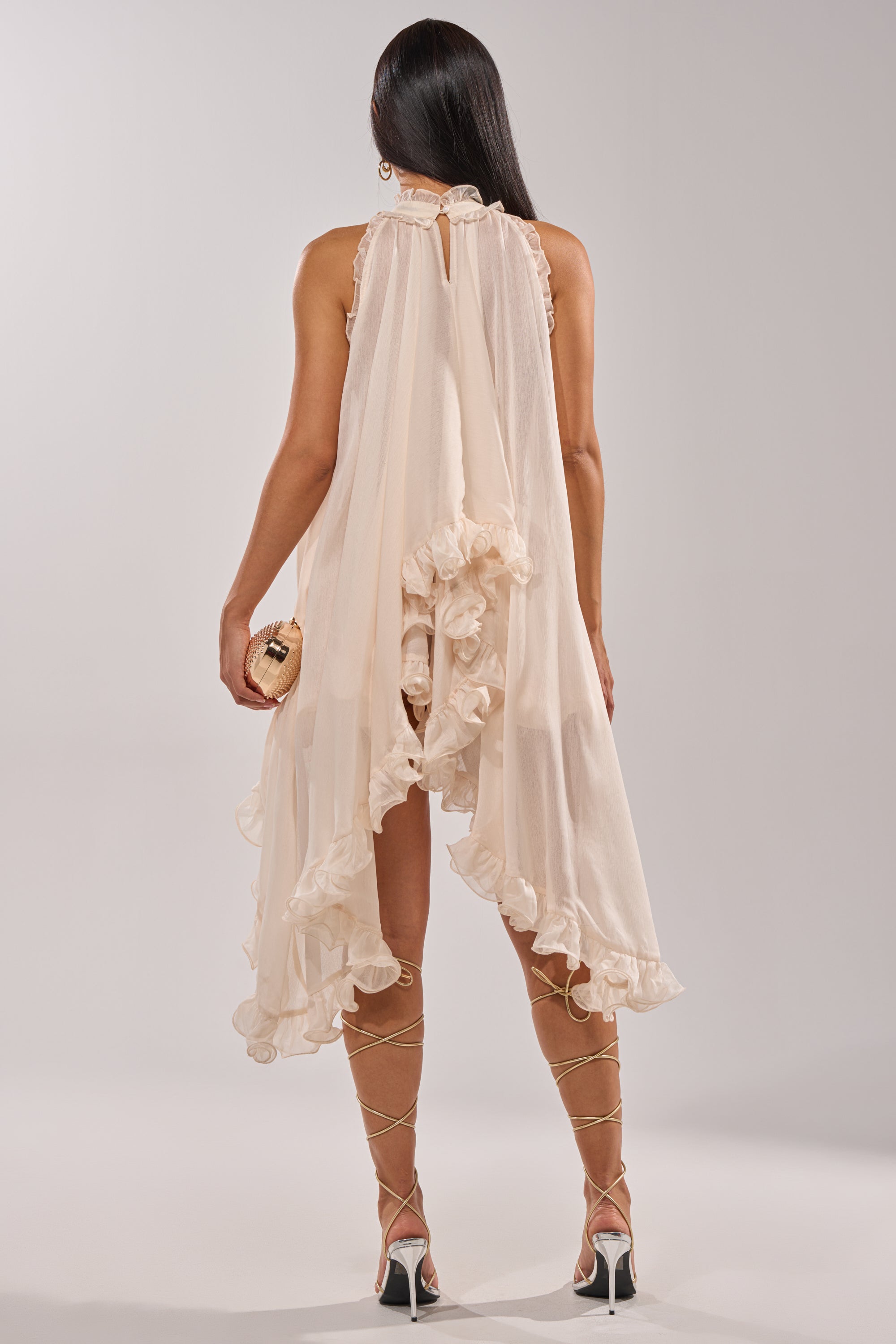 A woman with long straight hair faces away, wearing the TALK TO ME HIGH LOW MESH BLOUSE in cream as a romantic layer, paired with strappy heels and a small clutch purse against a plain light background. 7