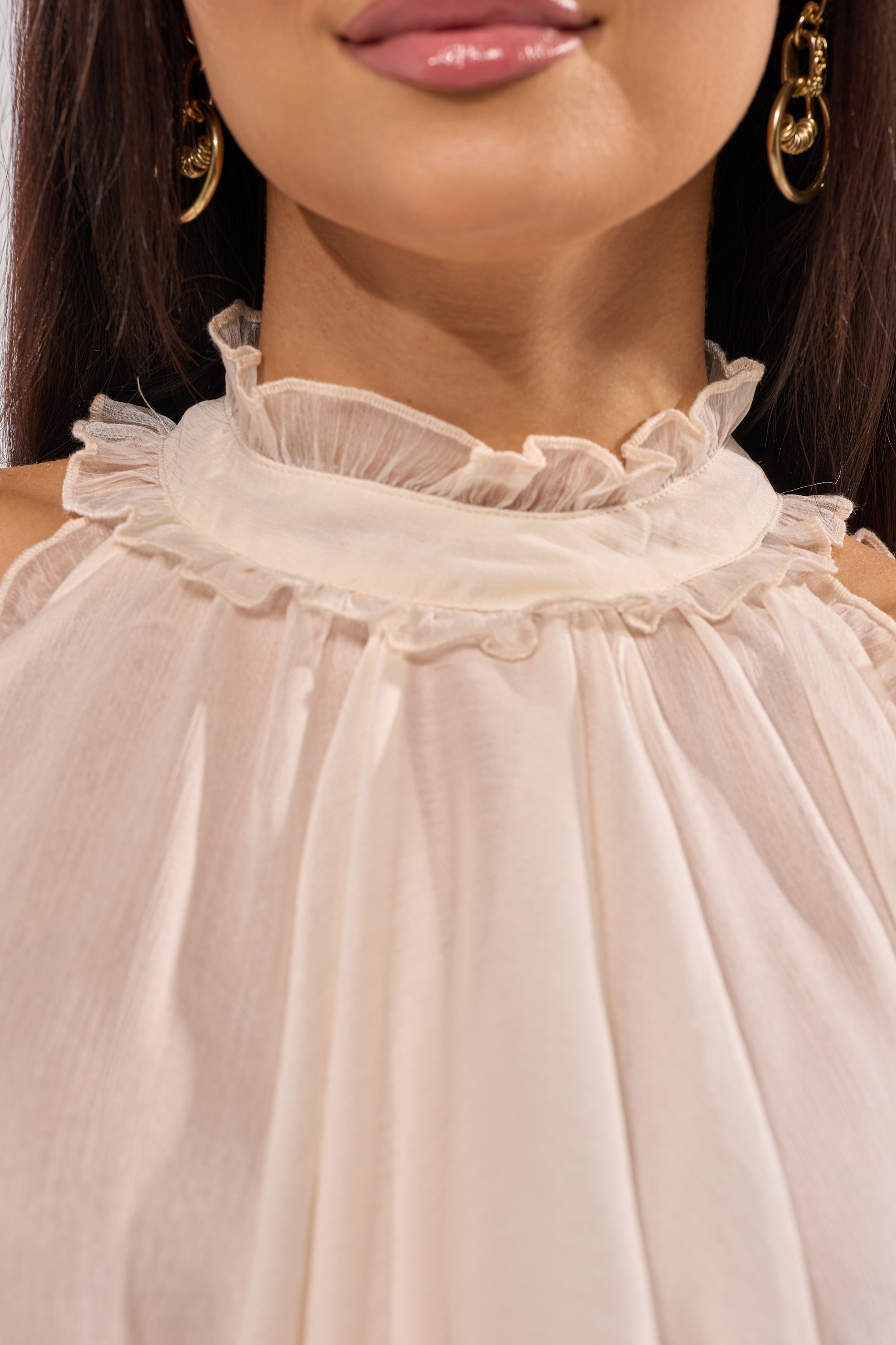 A woman wears gold hoop earrings and the TALK TO ME HIGH LOW MESH BLOUSE—an ivory, ruffled chiffon top with a sheer, high-neck design ideal for romantic layering. Only her lower face, neck, and upper chest are visible. 8