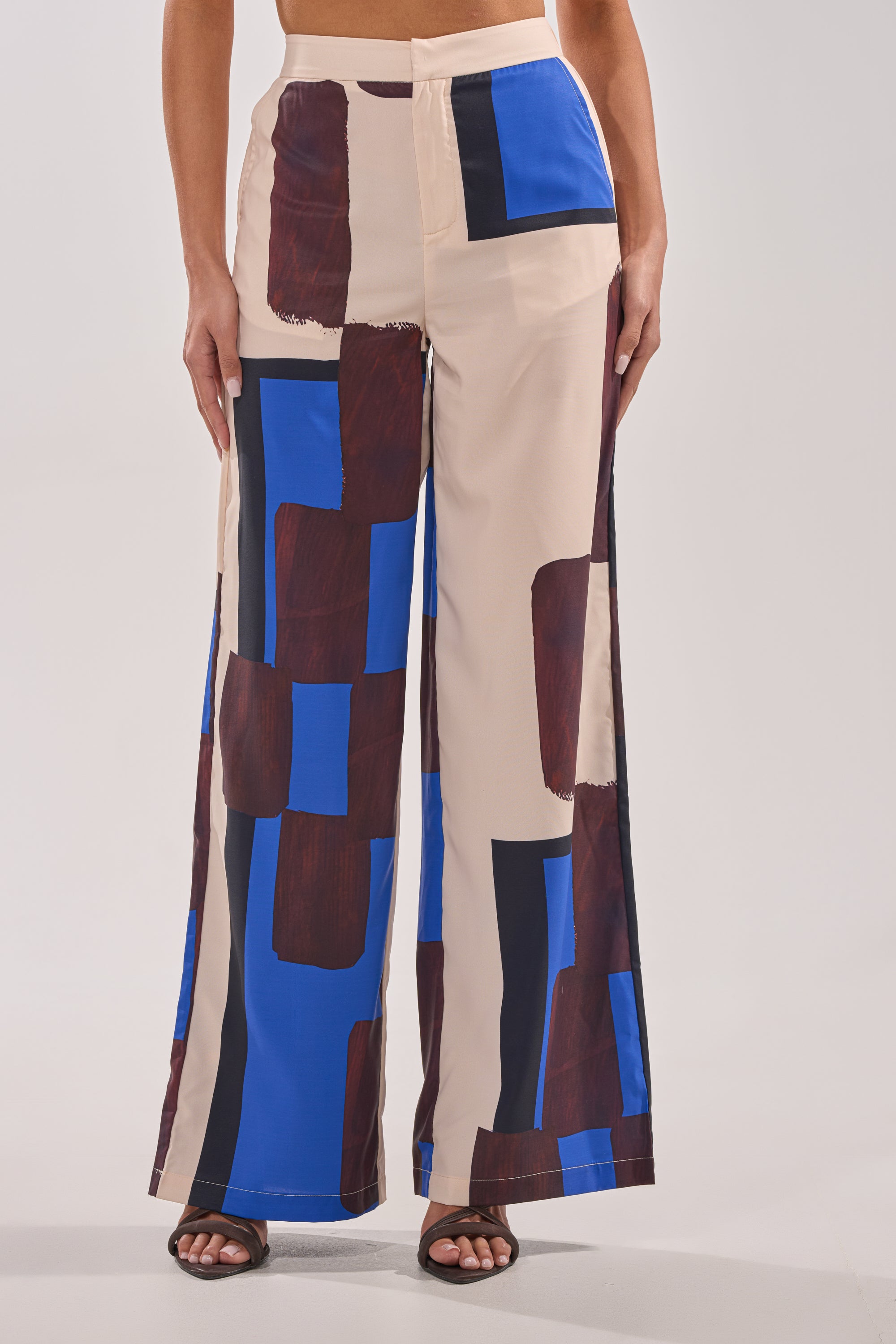 A person wears the ISLAND TIME TROUSER—high-rise, wide-leg pants with a beige base and abstract blue, black, and brown square patterns—standing slightly apart and paired with black open-toe heels. 1