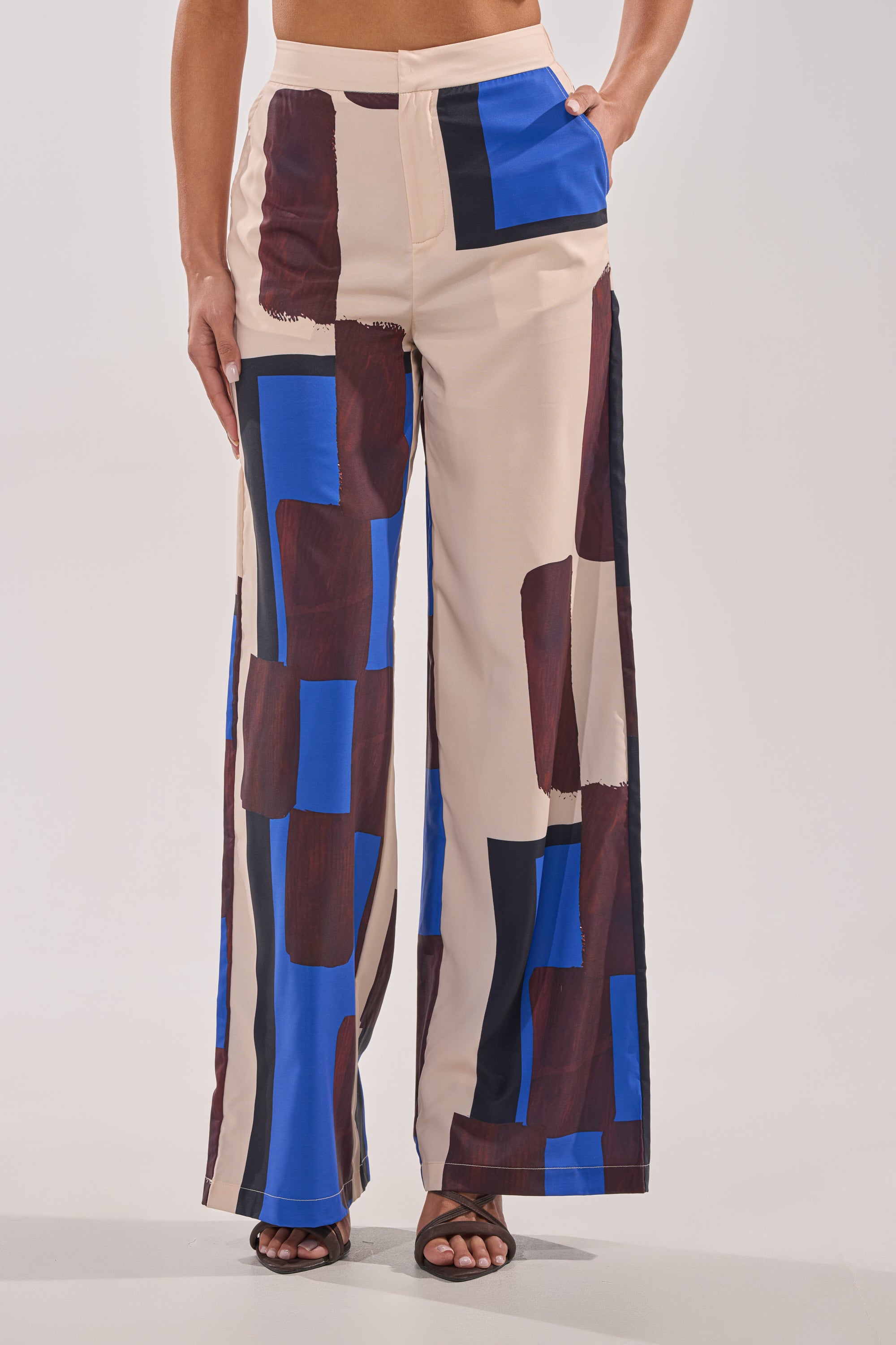 A person wears the ISLAND TIME TROUSER, featuring a cream satin base with bold brown, blue, and black geometric patterns, high-rise waist, and pockets. The look is paired with black open-toe heels. 2