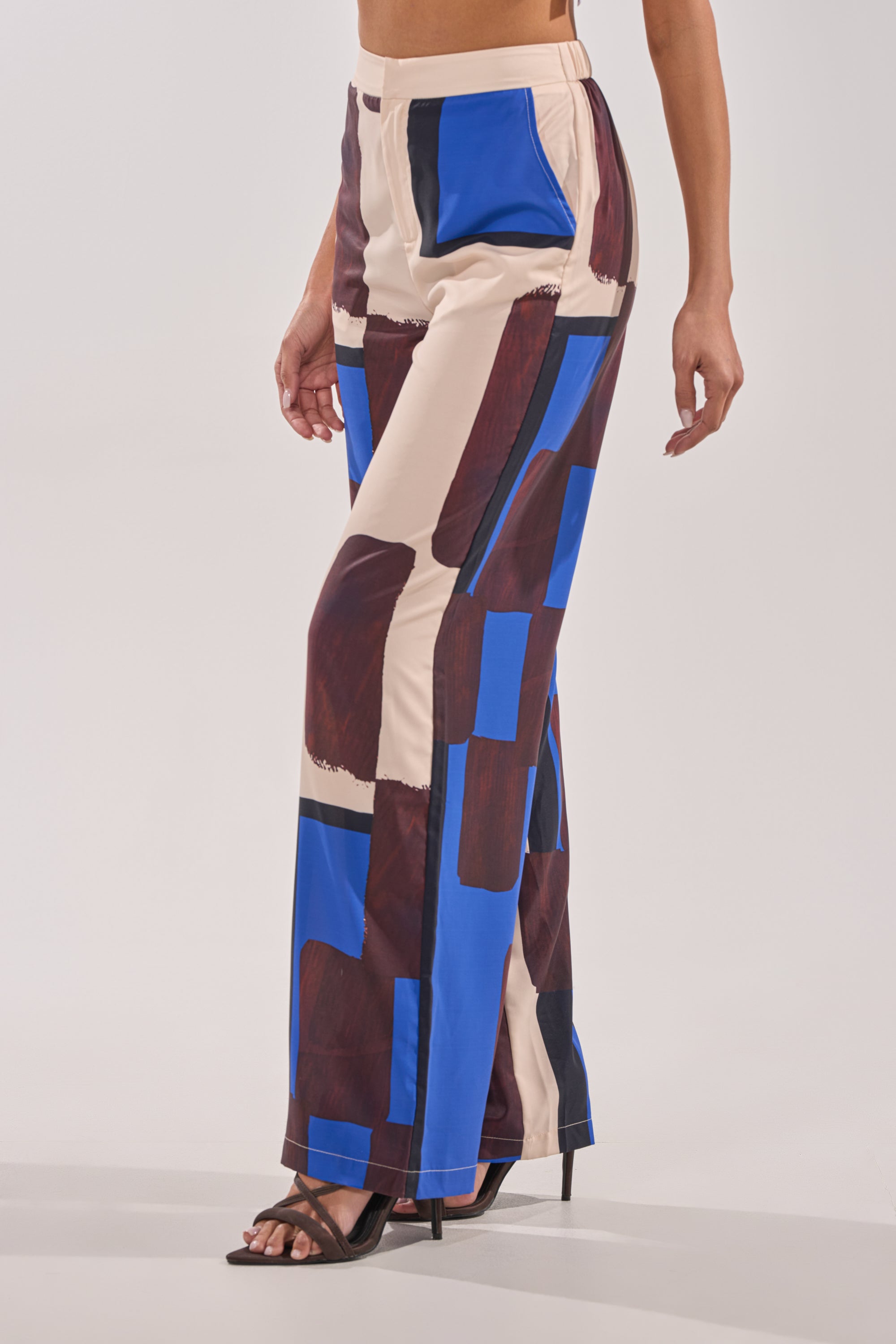 A person wearing the ISLAND TIME TROUSER featuring an abstract print in beige, blue, and brown with a high-rise waist pairs it with black high-heeled sandals, standing against a plain light background. 3