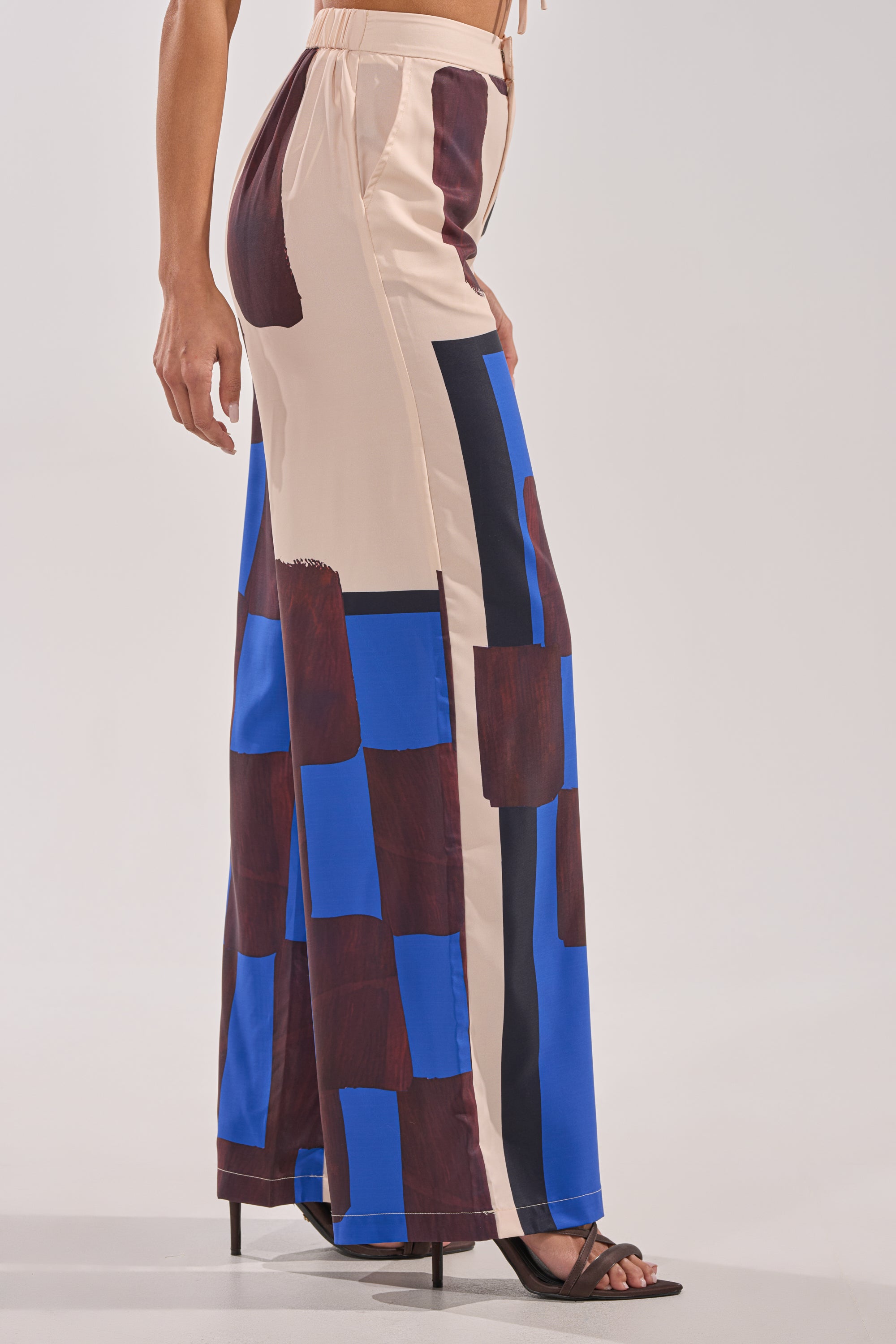 A person wearing the ISLAND TIME TROUSER—high-waisted pants with a beige base and bold brown and blue abstract blocks—paired with black heeled sandals. Only the lower half is visible, highlighting these chic print trousers. 4
