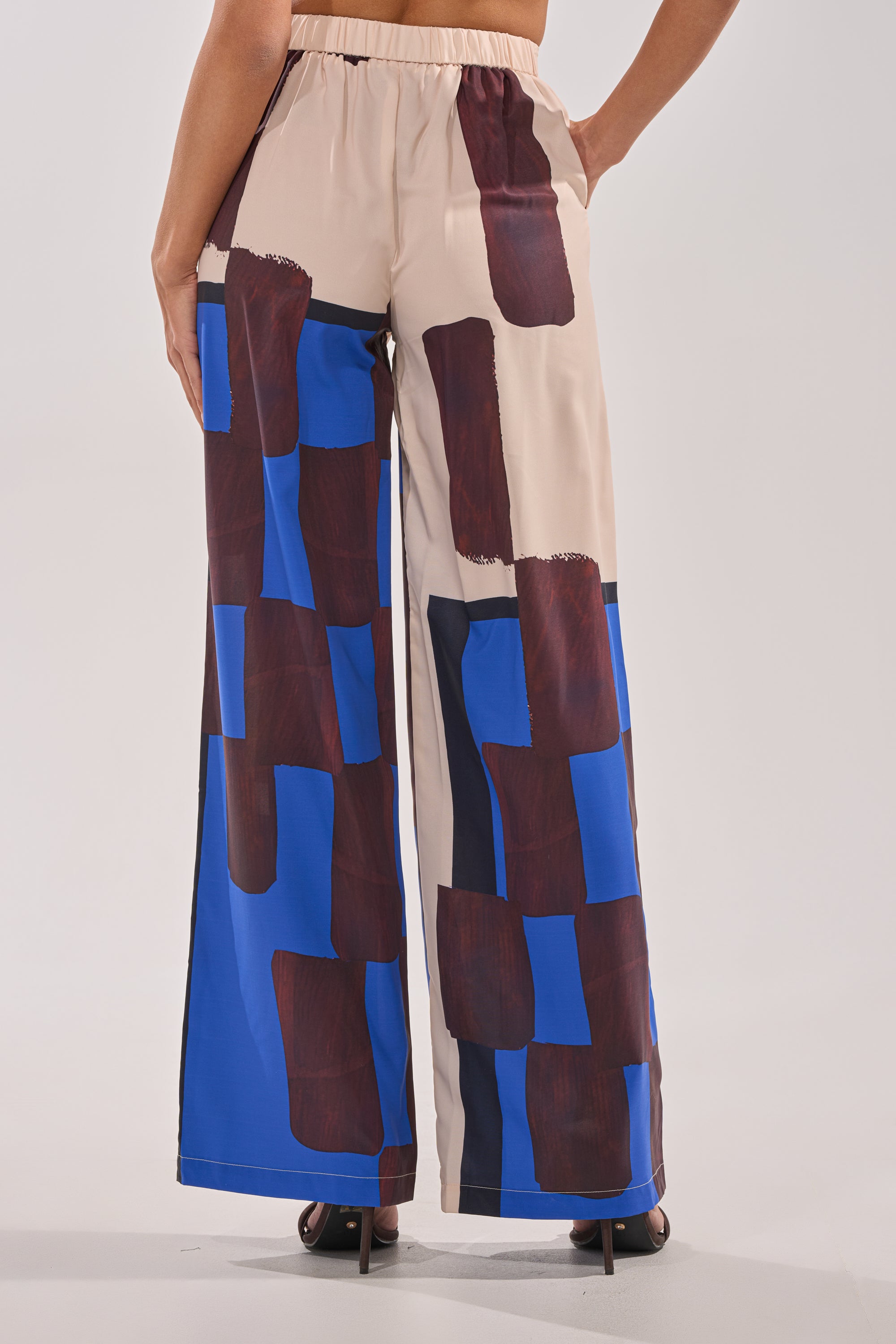 A person is seen from the back wearing the ISLAND TIME TROUSER, featuring a high-rise waist and wide-leg fit with an elastic waistband. The trousers showcase an abstract print in beige, blue, and dark brown rectangles. 5