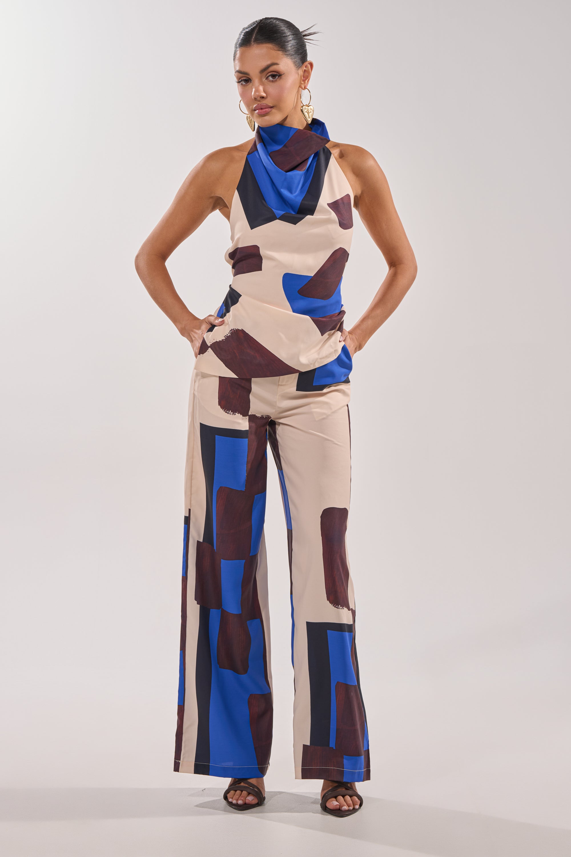 A woman poses confidently in the ISLAND TIME TROUSER, featuring a bold blue and dark brown abstract print, paired with a sleeveless beige top, large hoop earrings, and black open-toe heels. 7