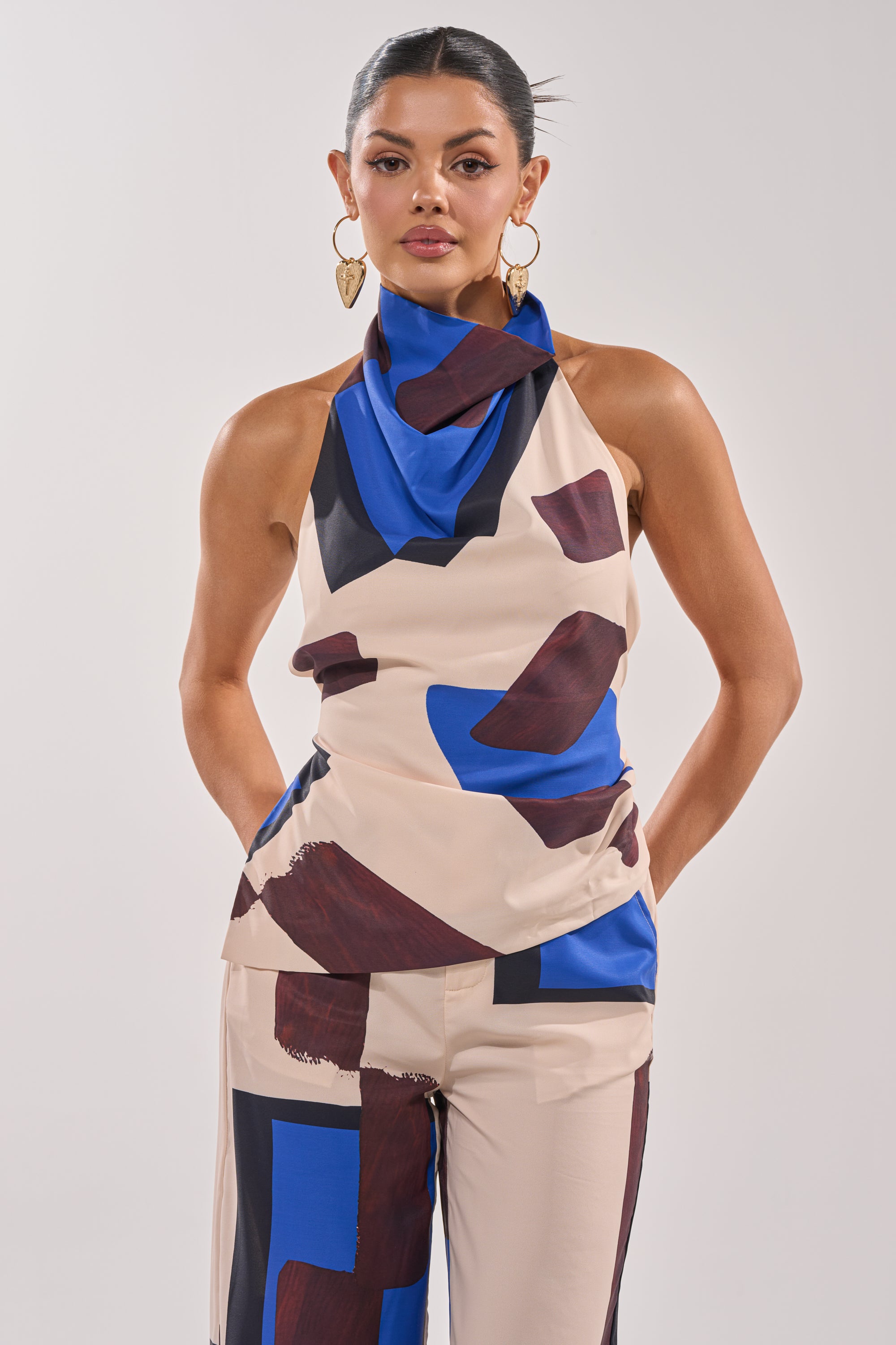 A woman stands facing forward, wearing the ISLAND TIME HALTER NECK, a sleeveless blouse with blue and brown abstract patterns. Her hands are in her pockets, hair pulled back, and she completes the look with large gold heart-shaped earrings. 0