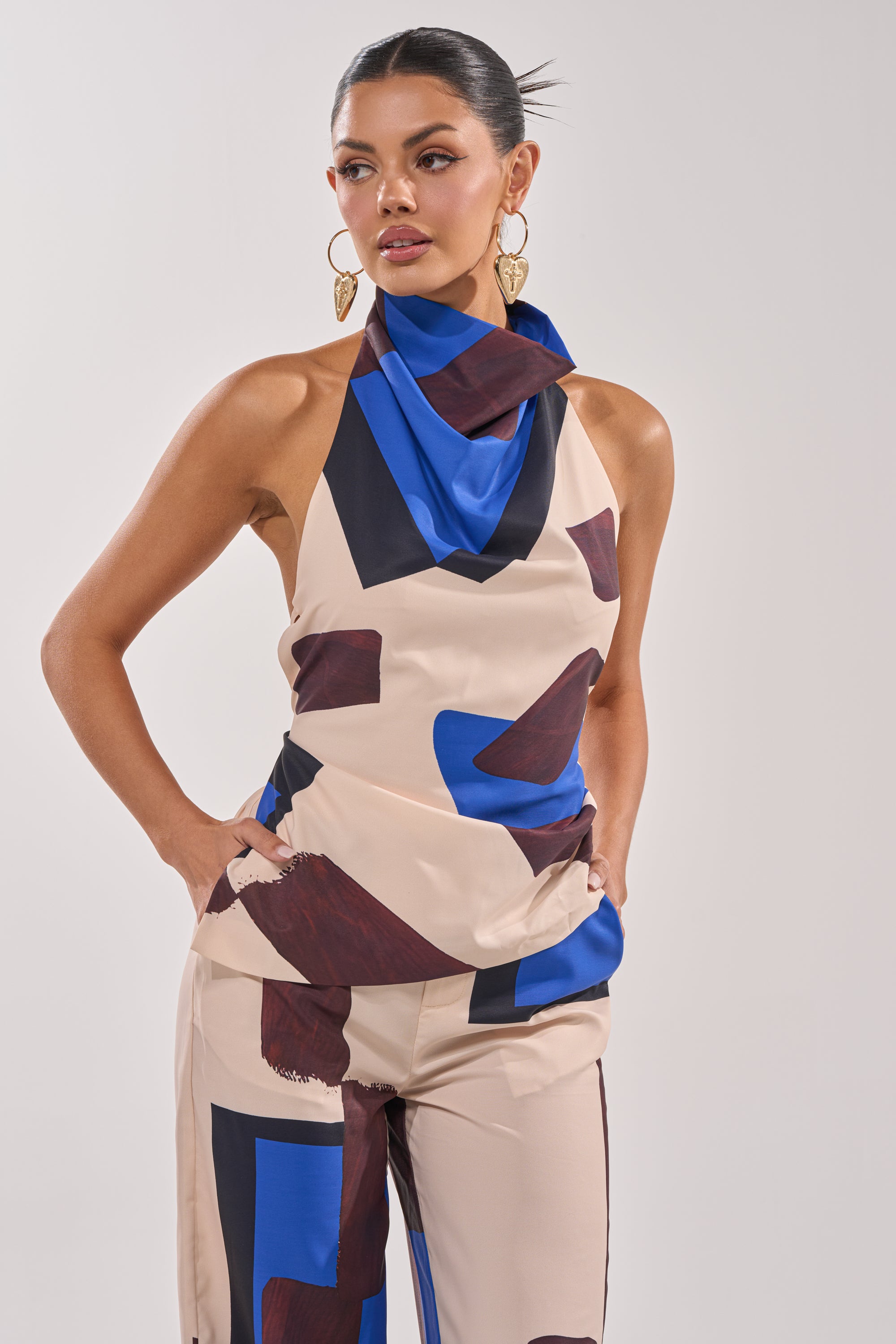 A woman models the ISLAND TIME HALTER NECK, featuring bold blue and maroon abstract prints. She accessorizes with large gold heart-shaped earrings and keeps her dark hair pulled back for a chic, modern look. 1