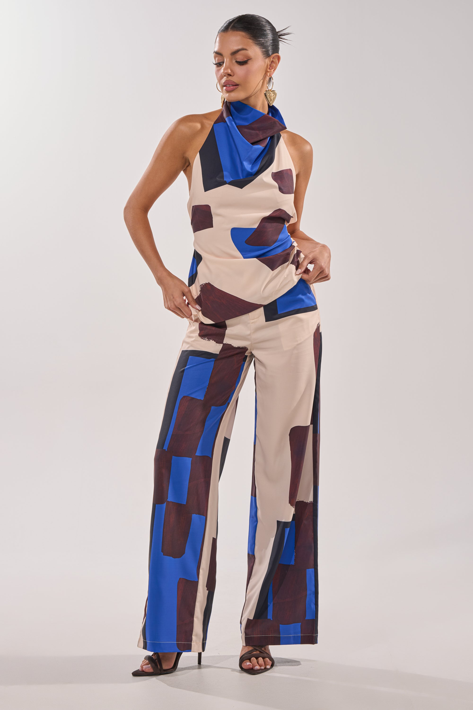 A woman wears the ISLAND TIME HALTER NECK, an open-back sleeveless top with matching wide-leg pants in a beige, blue, and brown geometric pattern. She pairs it with black heels, gold hoop earrings, and has her hair pulled back. 5