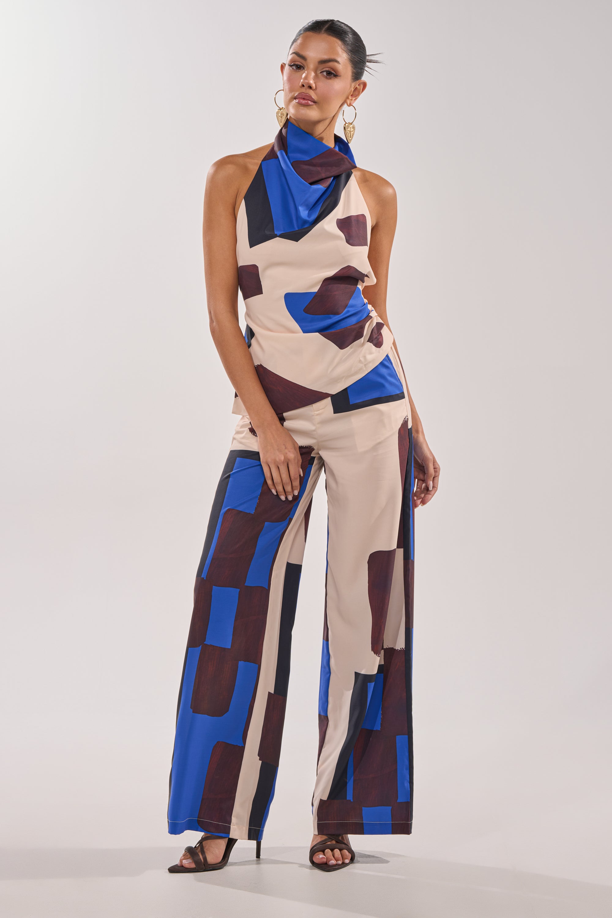 A woman stands wearing the ISLAND TIME HALTER NECK, a sleeveless draped top, paired with wide-leg pants featuring a bold geometric print in beige, blue, and brown. She accessorizes with hoop earrings, strappy black heels, and has her hair tied back. 6