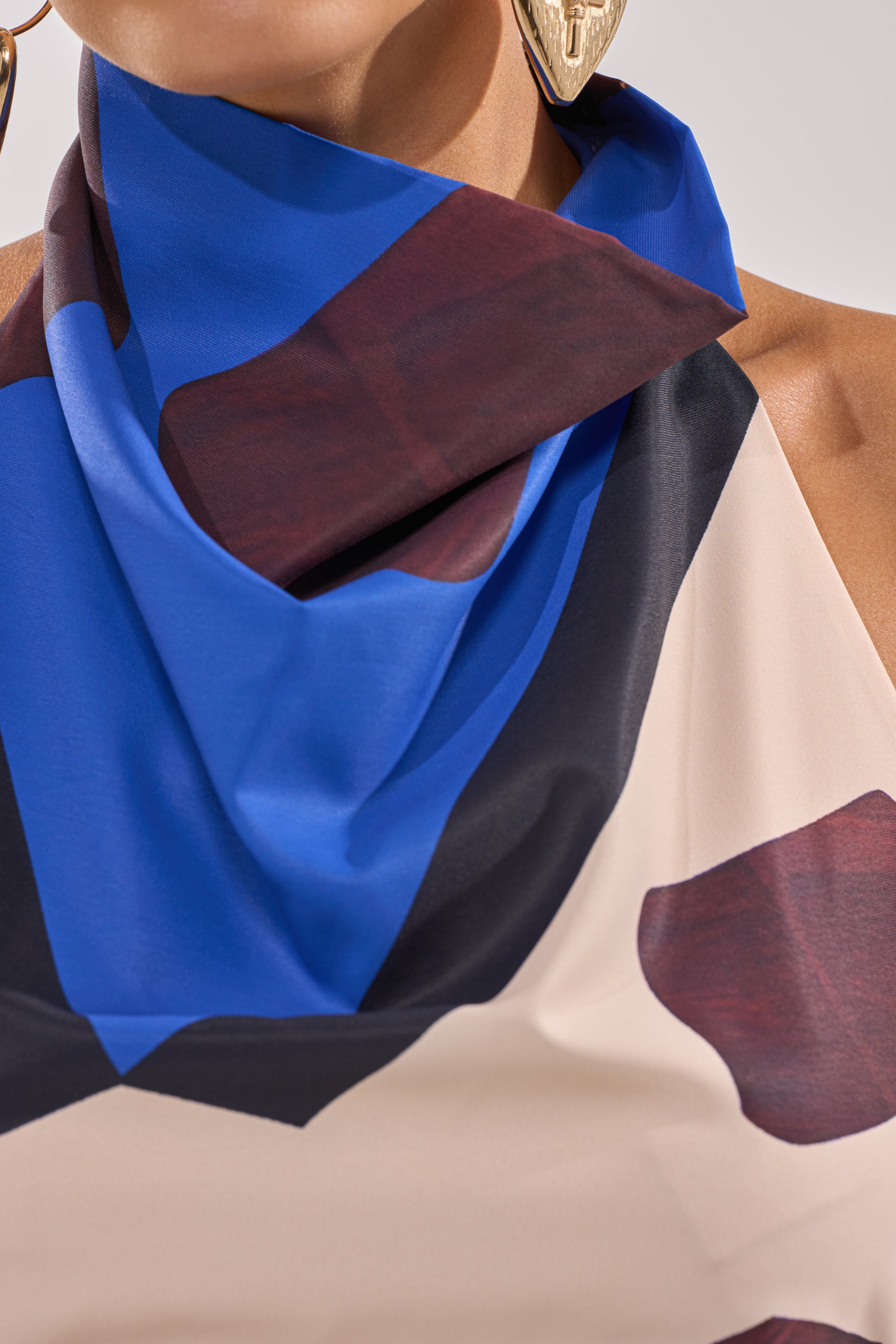 A close-up of someone wearing the ISLAND TIME HALTER NECK, a sleeveless draped top with bold blue, black, and burgundy geometric patterns and a matching scarf, paired with large gold face-shaped earrings. 8