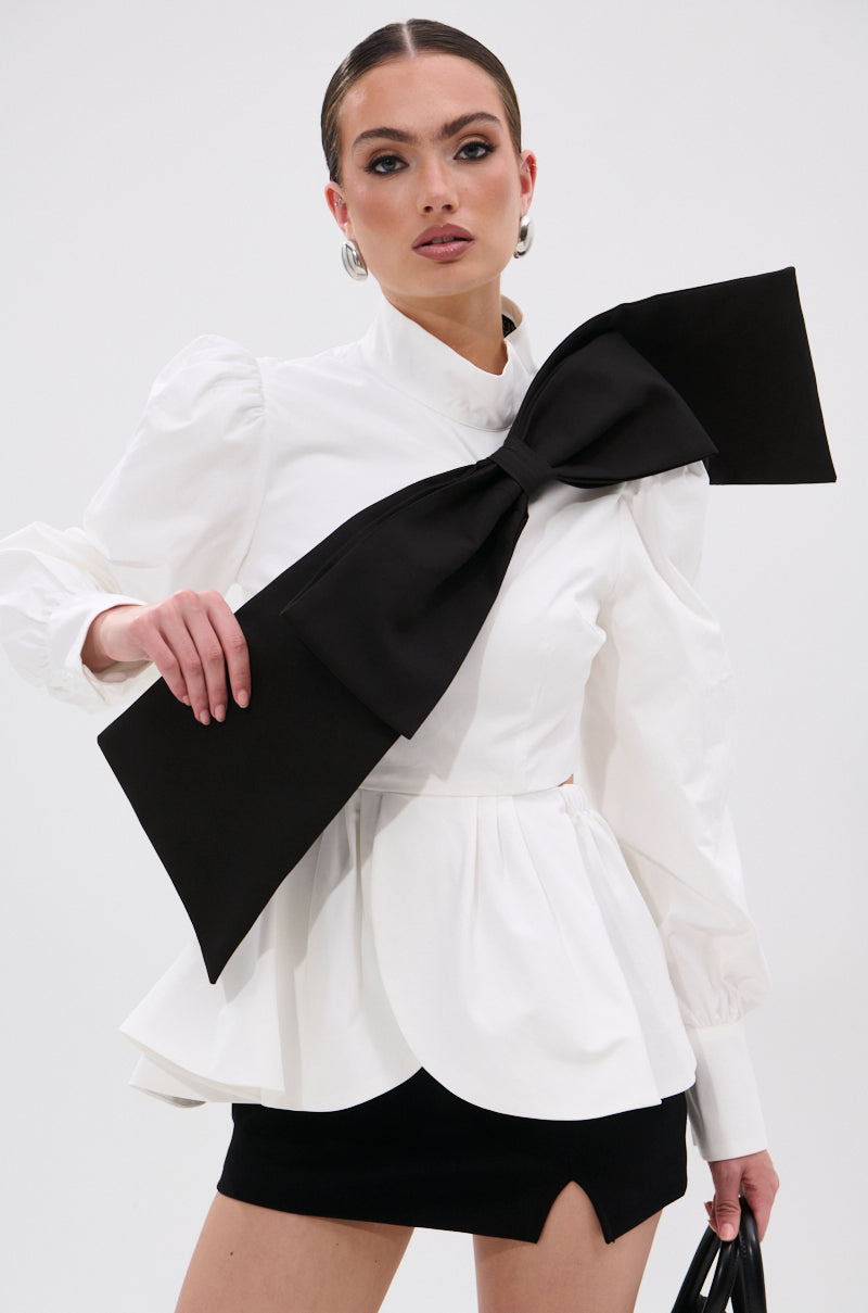 A woman confidently poses against a plain white background in the SO CHIC BOW BLOUSE, featuring a mock neckline and oversized black bow, paired with a short black skirt with a front slit. 0
