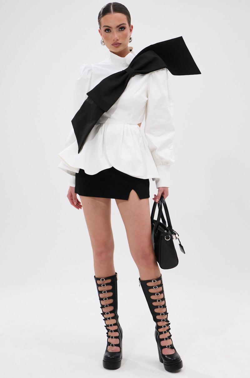 A woman stands against a white background wearing the SO CHIC BOW BLOUSE featuring a mock neckline and oversized black bow, paired with a short black skirt, knee-high lace-up boots, and holding a black handbag. 9