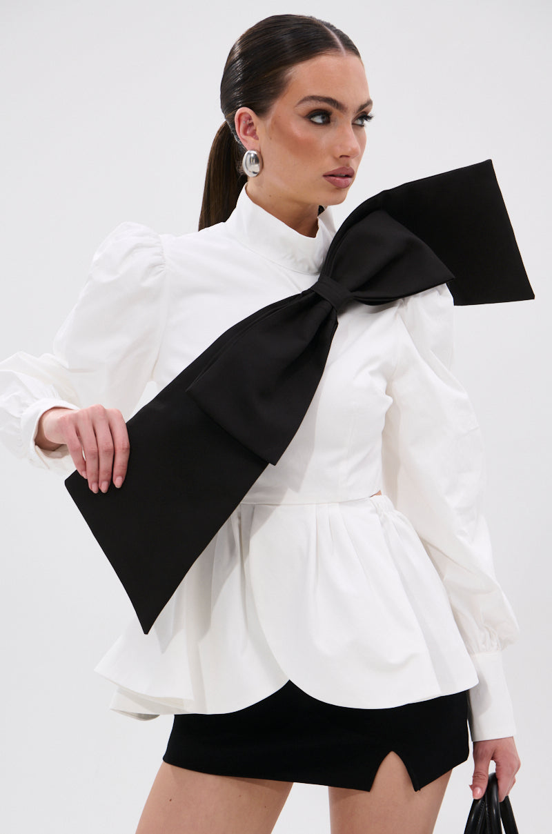A woman wears the SO CHIC BOW BLOUSE, a white top with puffed sleeves, mock neck, and an oversized black bow, paired with a short black slit skirt. She finishes the look with sleek hair and bold silver earrings. 1
