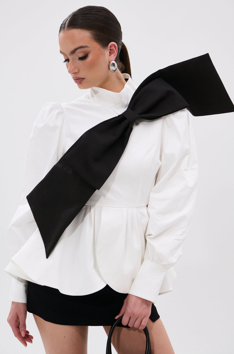 A woman wears the SO CHIC BOW BLOUSE with its mock neckline and oversized black bow across her shoulder and chest, paired with a black skirt, statement earrings, slicked-back hair, and a black handbag. 2