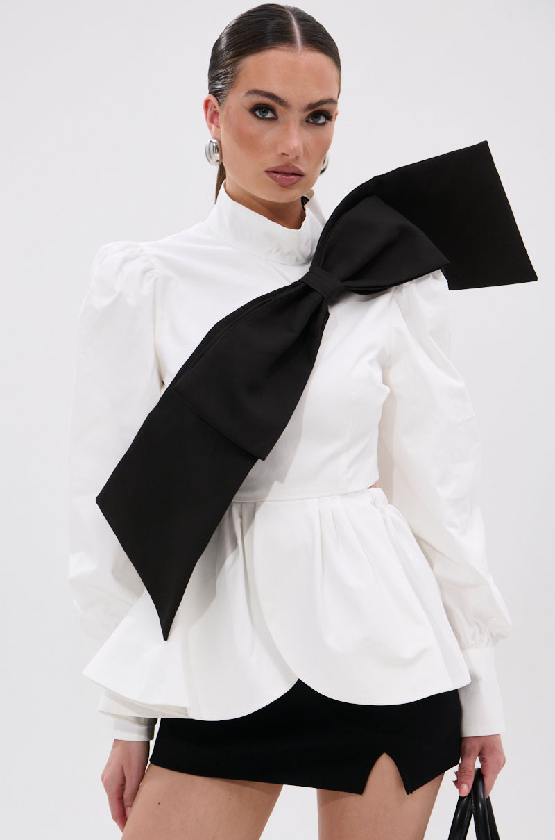 A woman models the SO CHIC BOW BLOUSE with puffed shoulders, mock neckline, and an oversized black bow on the chest, paired with a short black skirt with a front slit, standing against a plain light background. 3
