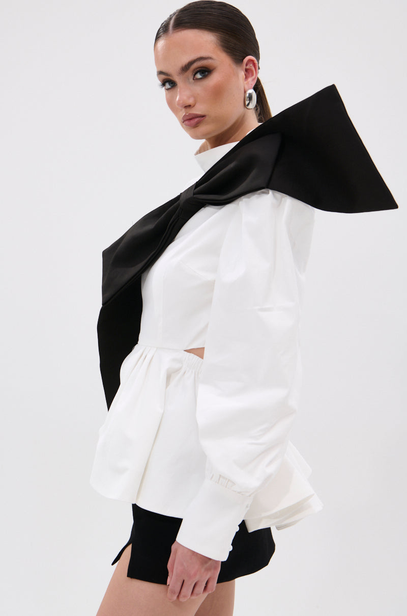 A woman models the SO CHIC BOW BLOUSE, showcasing its white peplum design, voluminous sleeves, mock neckline, and oversized black bow. She complements it with black shorts, slicked-back hair, and bold silver hoop earrings. 4