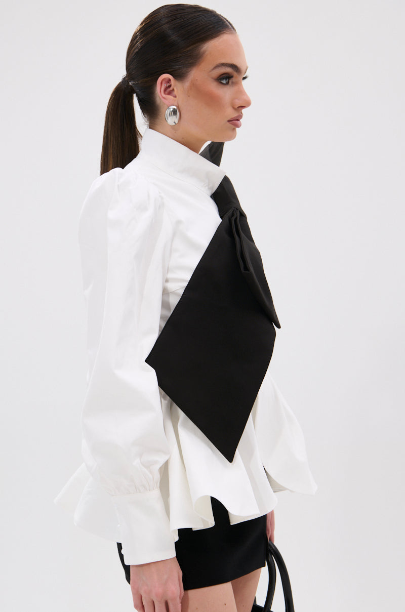 A woman with a sleek ponytail wears the SO CHIC BOW BLOUSE, designed with puffed shoulders, a mock neckline, and an oversized black bow, styled with a black mini skirt, statement silver earrings, and a chic black handbag. 5