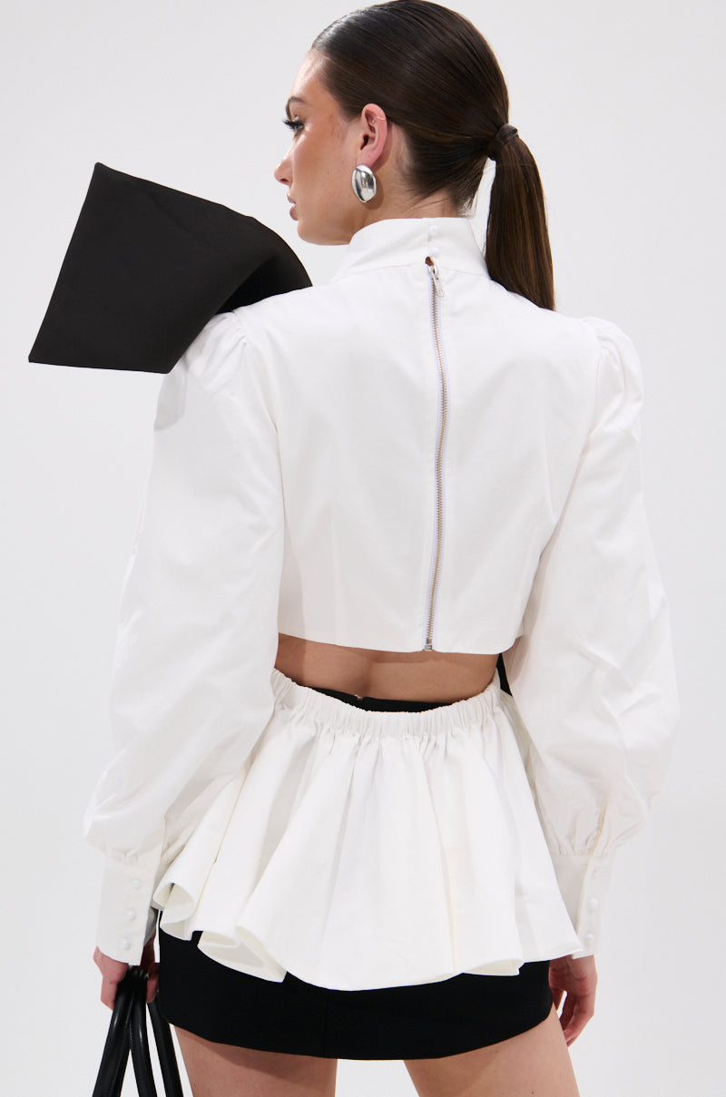 A woman with a sleek ponytail wears the SO CHIC BOW BLOUSE, a white long-sleeve top with a mock neckline, zipper back, bold black geometric shoulder detail, and ruffled cropped hem, paired with a black mini skirt. 6