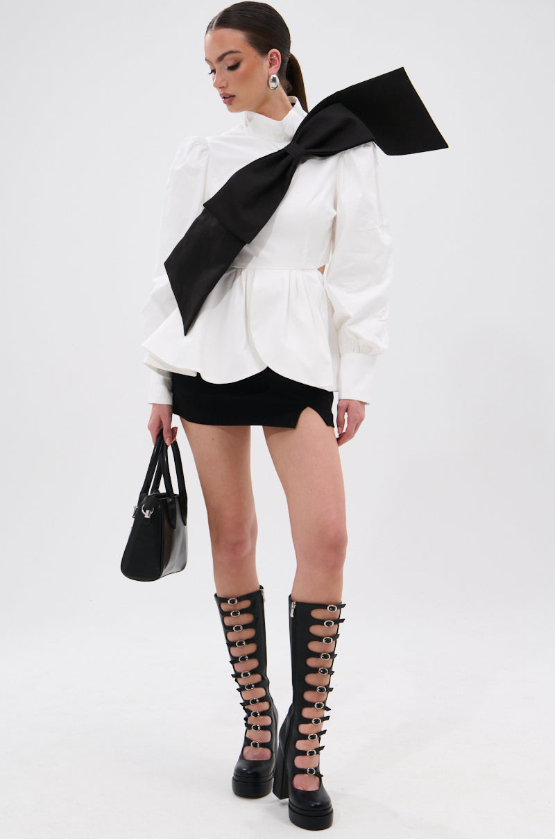 A woman in the SO CHIC BOW BLOUSE, featuring puffed sleeves, a mock neckline, and an oversized black bow, pairs it with a black mini skirt, knee-high lace-up boots, and a black handbag against a plain white background. 8