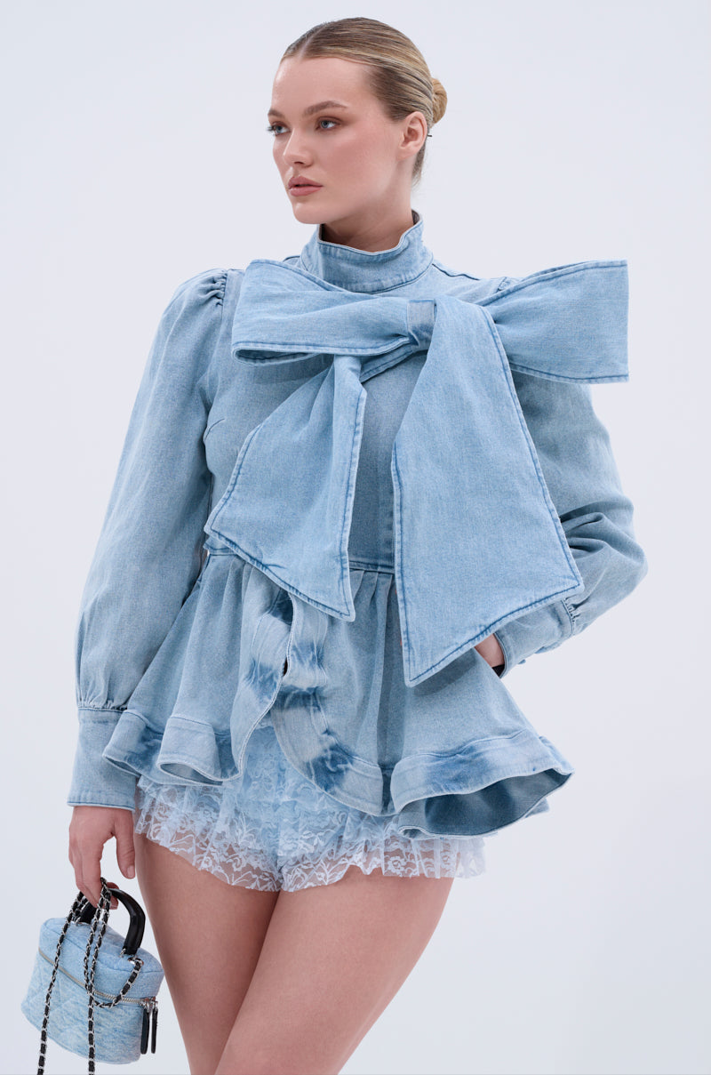 A woman poses against a plain light background wearing the PHOEBE DENIM BOW BLOUSE with puff sleeves and ruffled peplum hem, paired with lace shorts and holding a small chain-strap denim bag.