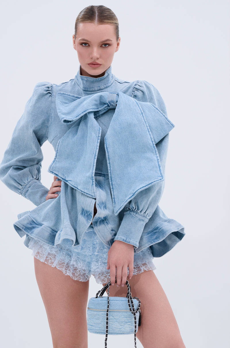 A woman poses in the PHOEBE DENIM BOW BLOUSE with puff sleeves, a ruffled peplum hem, lace-trimmed shorts, and a matching denim handbag against a plain light blue background.