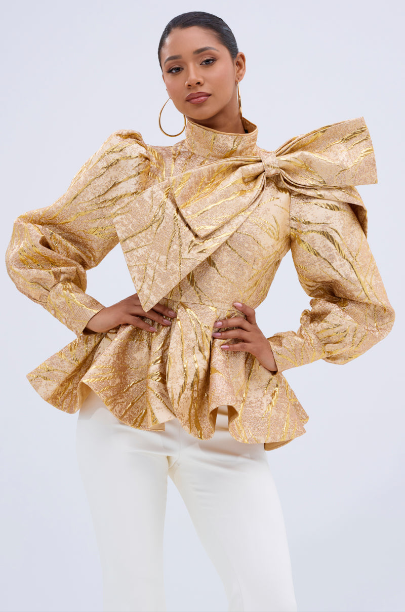 A woman exudes confidence in the LANA GOLDEN BLOUSE, featuring shiny gold jacquard with a statement shoulder bow, paired with white pants and large hoop earrings. Her hair is pulled back for a chic, polished finish. 0