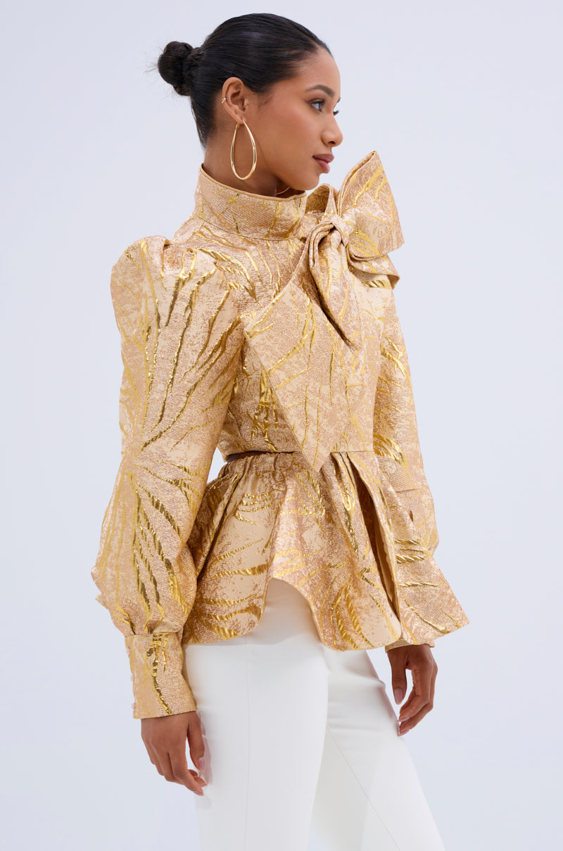 A woman in profile wears the LANA GOLDEN BLOUSE, a metallic jacquard top with puff sleeves and a bold shoulder bow, paired with white pants. Her hair is styled in a neat bun, complemented by large hoop earrings. 1