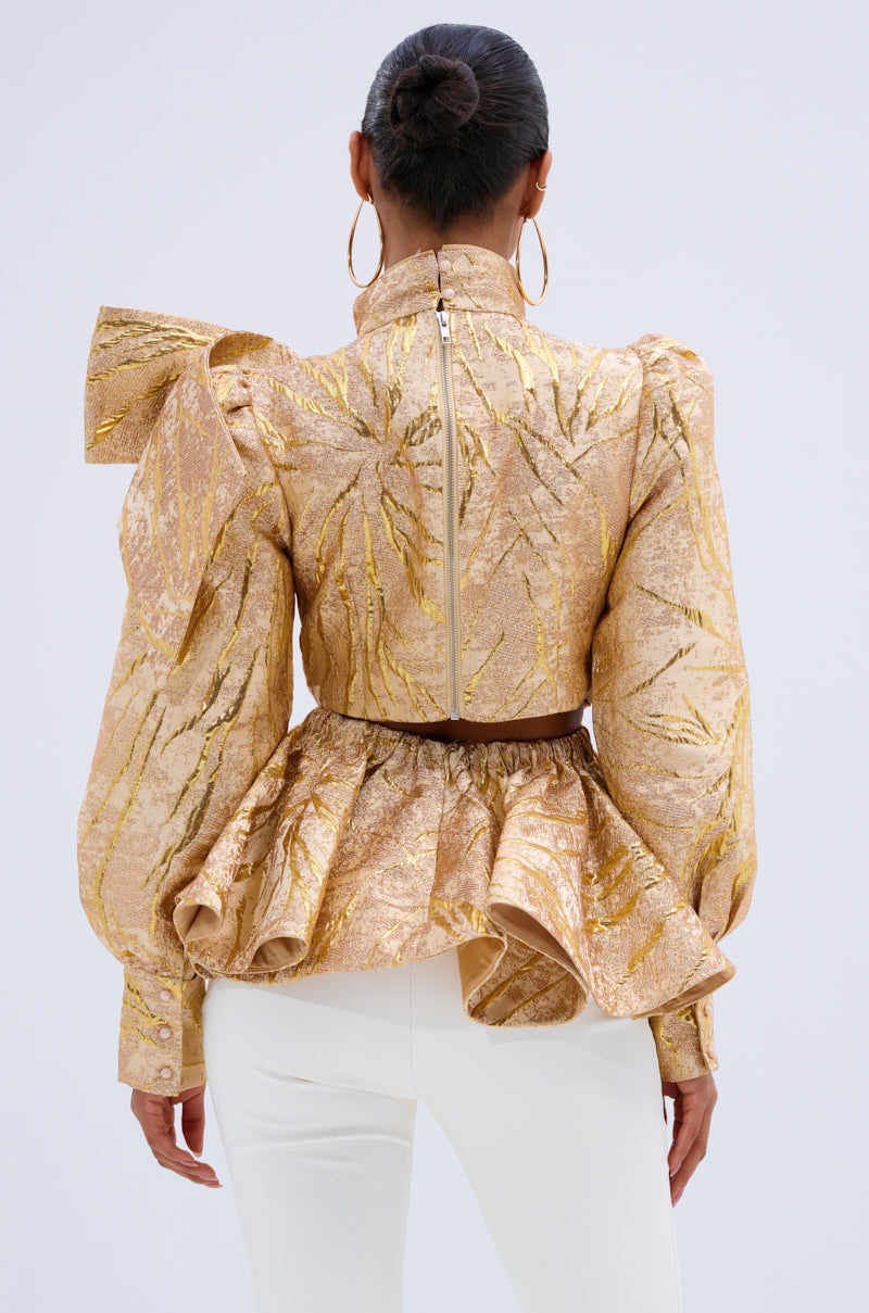 A woman is seen from the back wearing the LANA GOLDEN BLOUSE— a gold metallic jacquard top with puff sleeves and a peplum hem— paired with white fitted pants. Her hair is in a neat bun, accessorized with large hoop earrings. 3