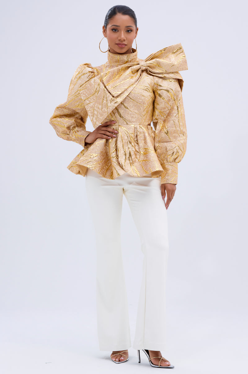 A woman wears the LANA GOLDEN BLOUSE, featuring metallic jacquard fabric, voluminous sleeves, and a statement bow on the shoulder, styled with white flared pants and open-toe heels against a simple light background. 4
