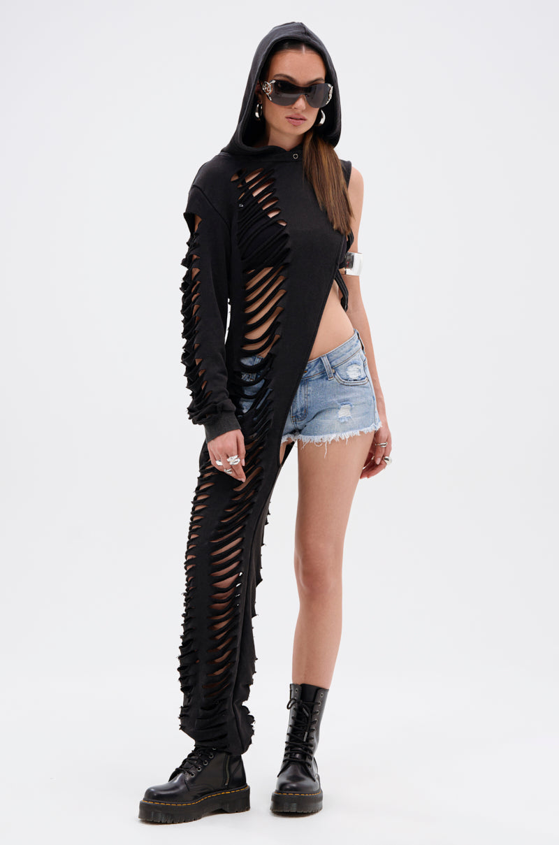 A woman stands against a white background wearing the ONE OF A KIND SWEATSHIRT—a black, heavily shredded, asymmetrical, oversized hooded top—paired with distressed denim shorts, black combat boots, sunglasses, and a metallic arm cuff.