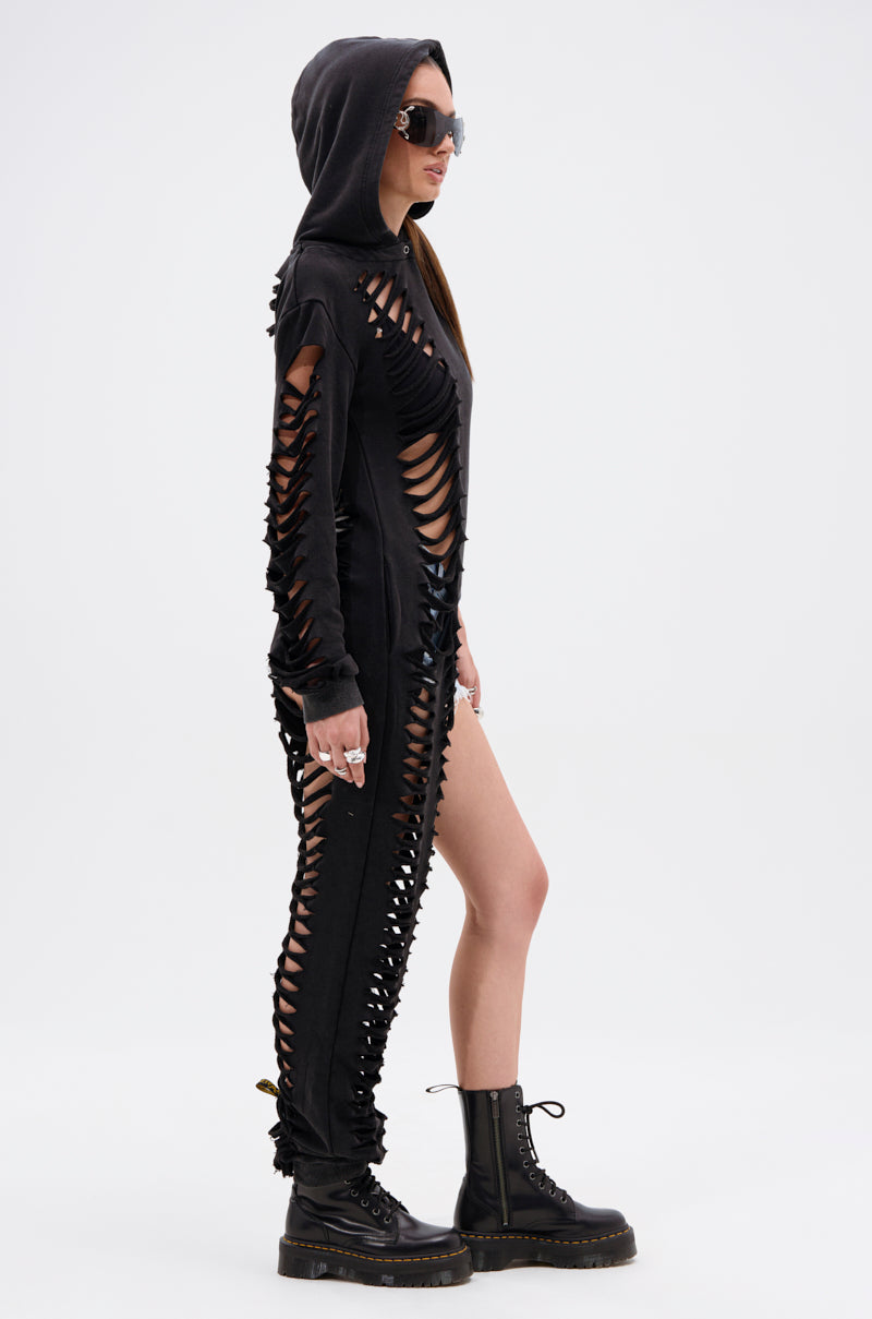 A person in the ONE OF A KIND SWEATSHIRT, a black hooded piece with an asymmetrical silhouette and shredded cutouts along the sides and arms, paired with black combat boots and sunglasses, stands in profile against a plain white background.