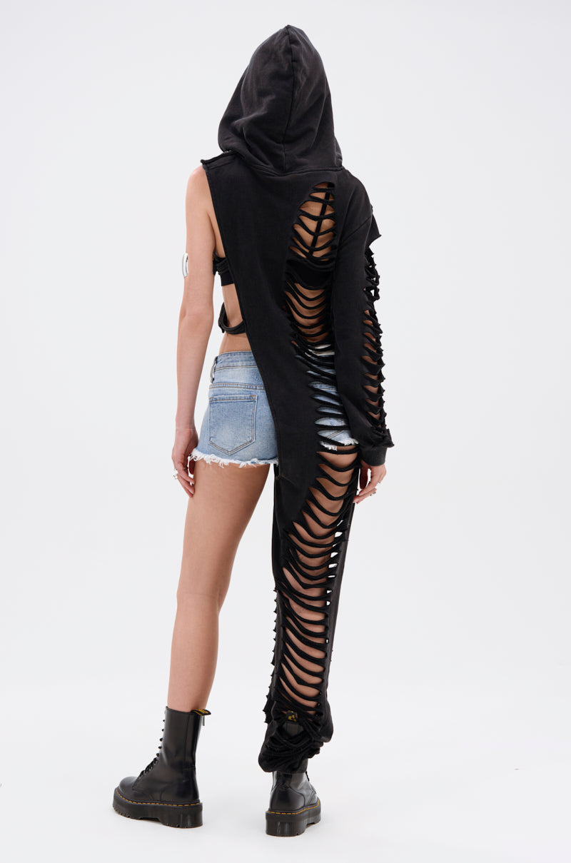 A person stands facing away, wearing black combat boots, denim shorts, and the ONE OF A KIND SWEATSHIRT—an oversized asymmetrical black hoodie with one sleeve and pant leg, both detailed with horizontal slits for a distressed look.