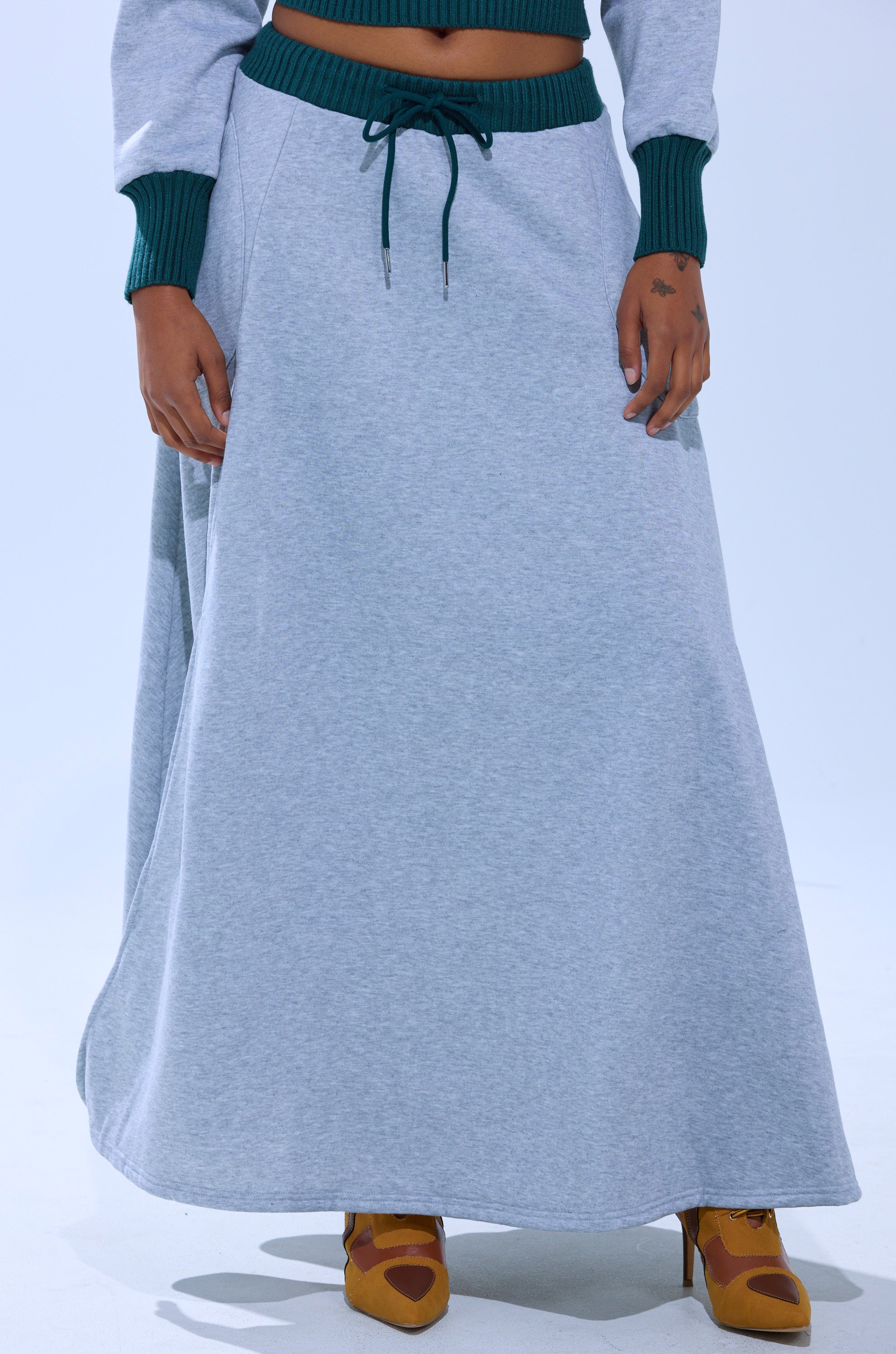 A person wears the COLLEGIATE FRENCH TERRY MAXI SKIRT—a long, light gray high-rise skirt with a dark green waistband and sweater cuffs—paired with mustard yellow heeled shoes. The image is cropped from the chest down.