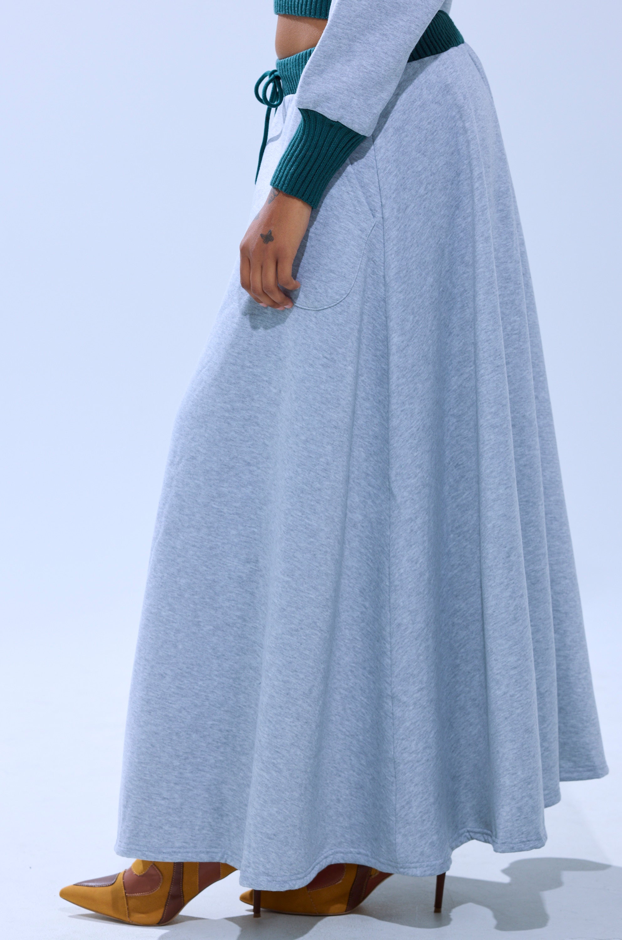 A person models the COLLEGIATE FRENCH TERRY MAXI SKIRT in light gray with a green top and brown heels against a plain background. The waist-down shot highlights the skirt's elegant, high-rise silhouette.