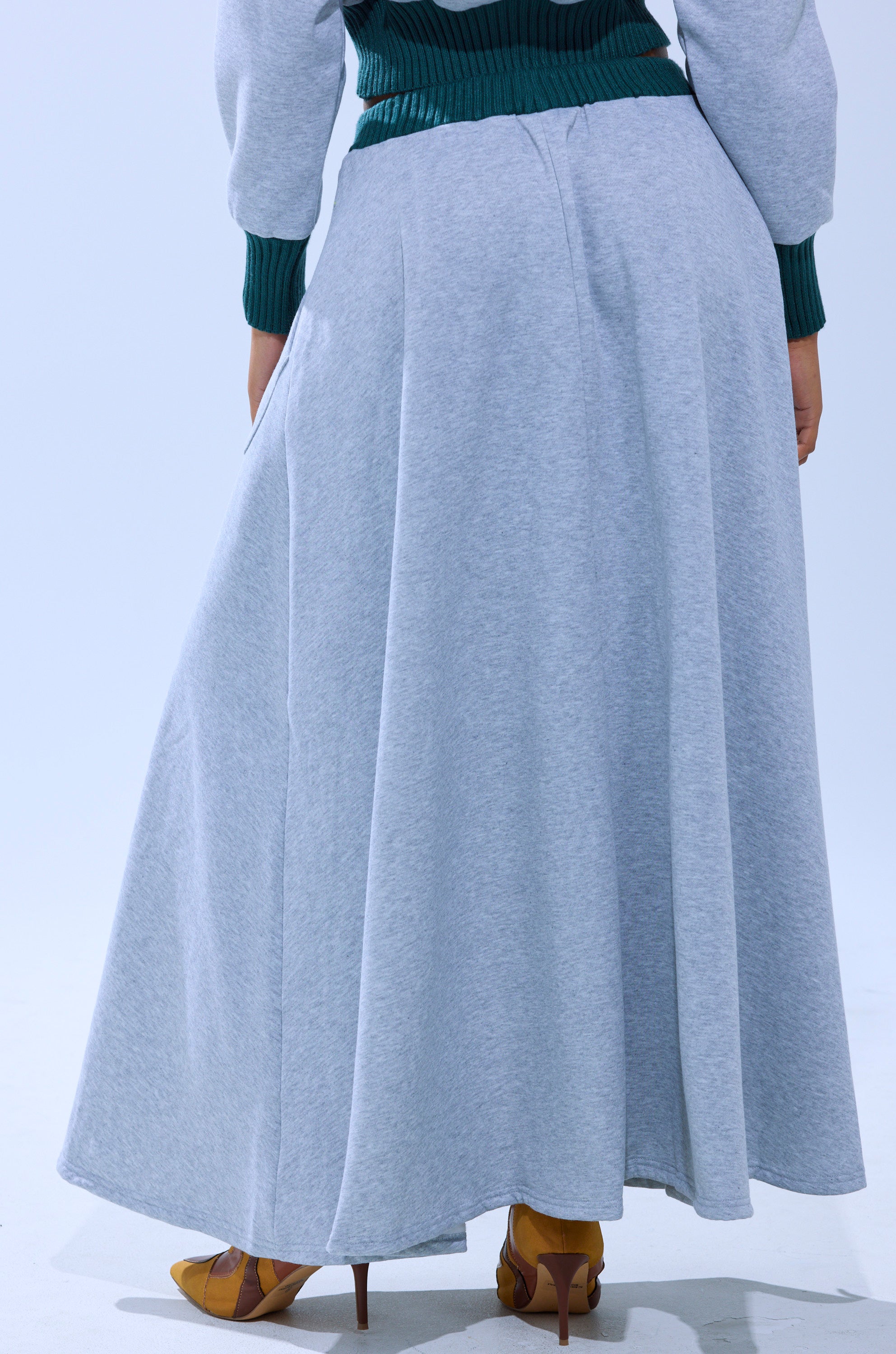 A person wearing the COLLEGIATE FRENCH TERRY MAXI SKIRT in light gray and mustard yellow heels faces away, with a green ribbed sweater partially visible. The background is plain and light-colored.