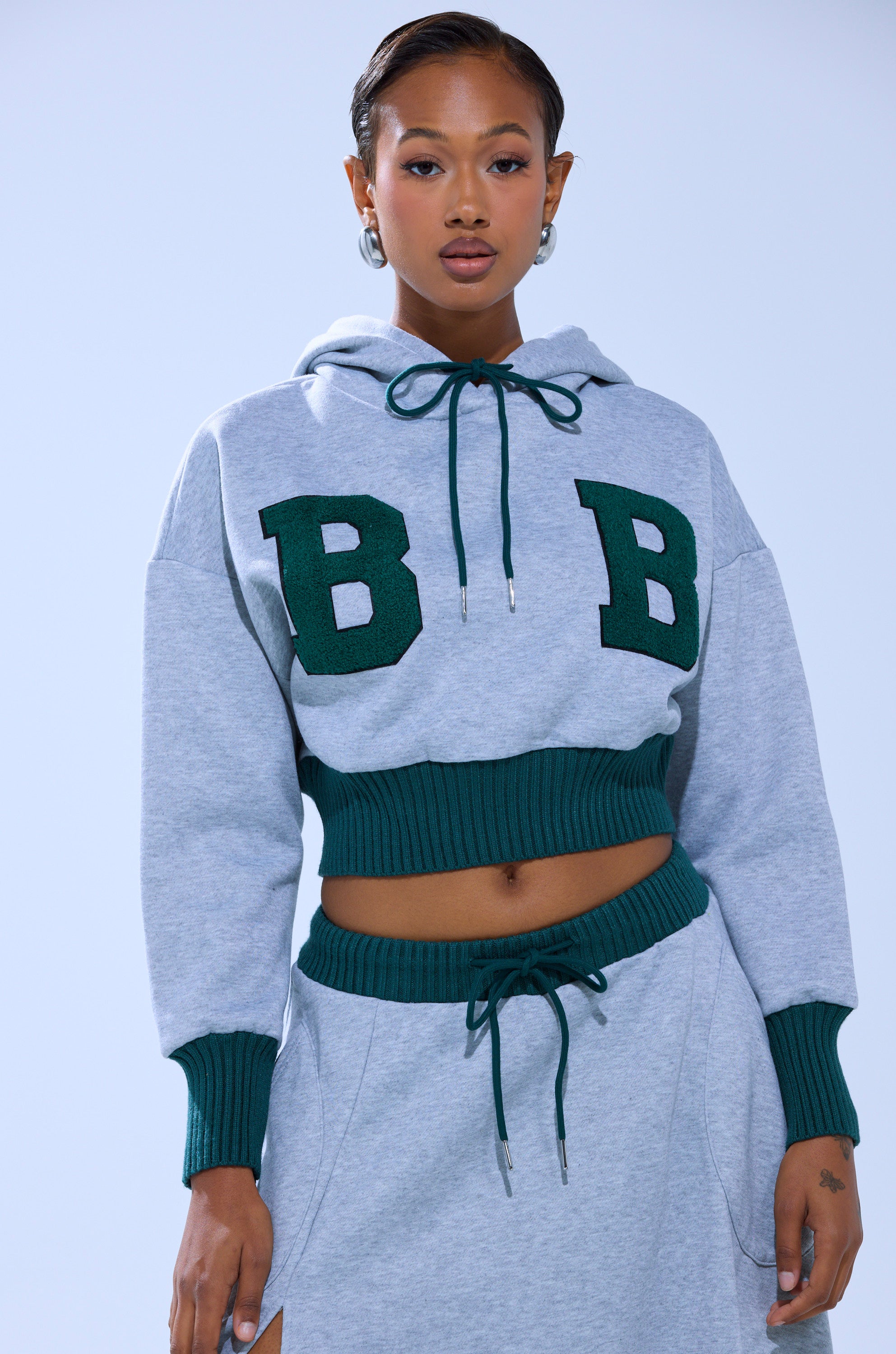 A woman with slicked-back hair and hoop earrings poses against a light blue background wearing the COLLEGIATE FRENCH TERRY HOODED SWEATSHIRT in gray with green “BB” letters, paired with matching gray sweatpants featuring green trim.